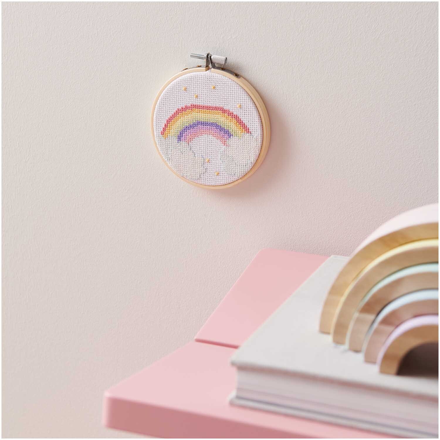 Kit de broderie Mini Kit Image Arc-en-ciel Ø10 cm from Rico Design Stickpackung Mini Kit Regenbogen Bild Ø10 cm zeigt einen bunten Regenbogen auf weißem Stoff, aufgehängt an einer rosa Wand.