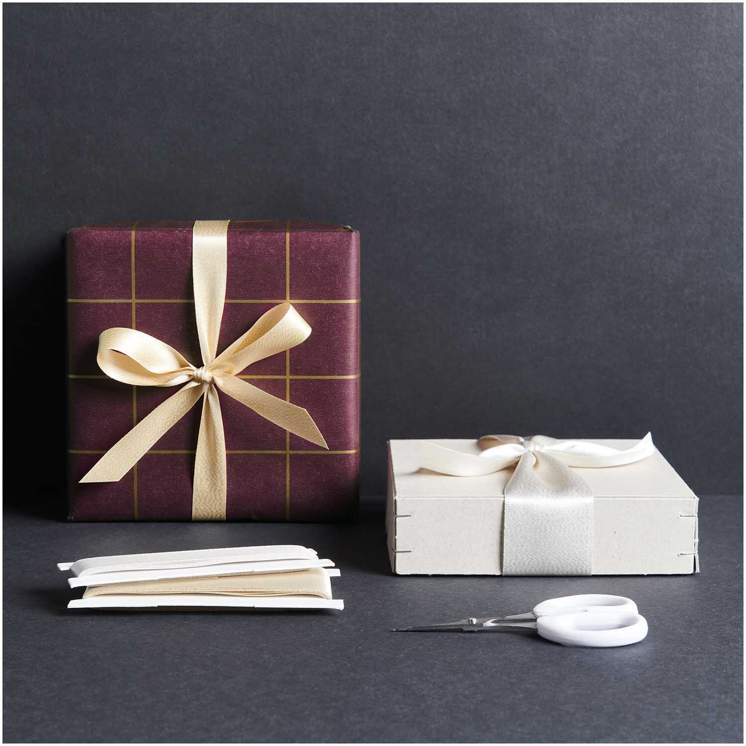 Paper Poetry Geschenkpapier Raster bordeaux-gold 70cm 2m 80g/m² von Rico Design Paper Poetry Geschenkpapier Raster bordeaux-gold umhüllt ein Geschenk mit goldener Schleife. Daneben ein weißes Paket mit Schleife auf dunklem Hintergrund.