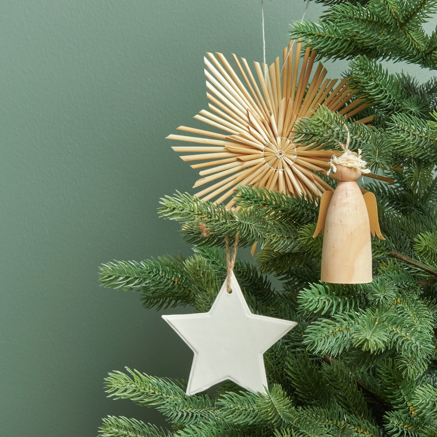 Holzengel mit Krone neben Strohstern und weißem Porzellanhänger in Tannenbaum von Rico Design.