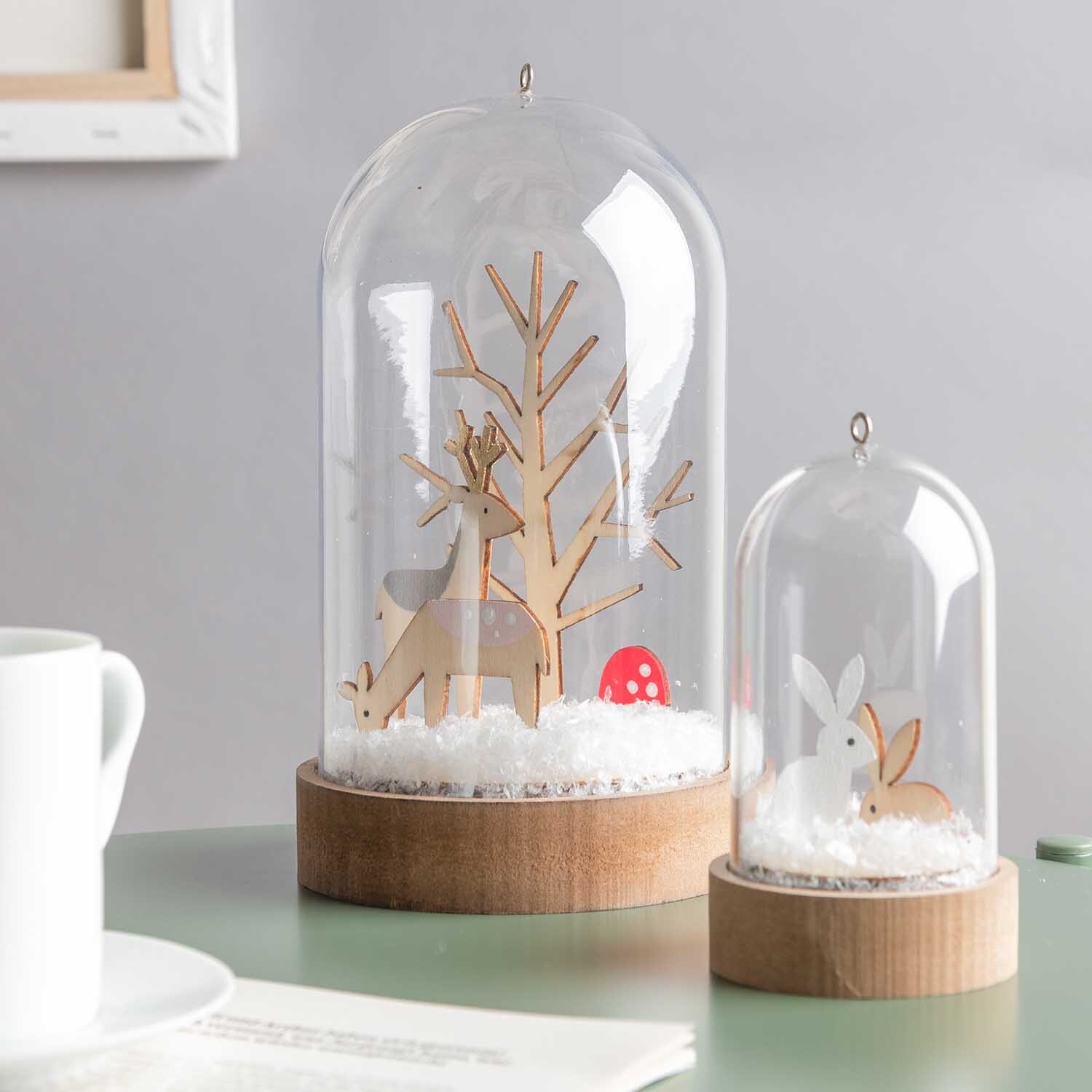 Dekoschnee 100g von Rico Design Dekoschnee 100g: Zwei Glasglocken mit Holzfiguren von Rehen und Hasen auf Dekoschnee, ideal für winterliche Dekorationen.