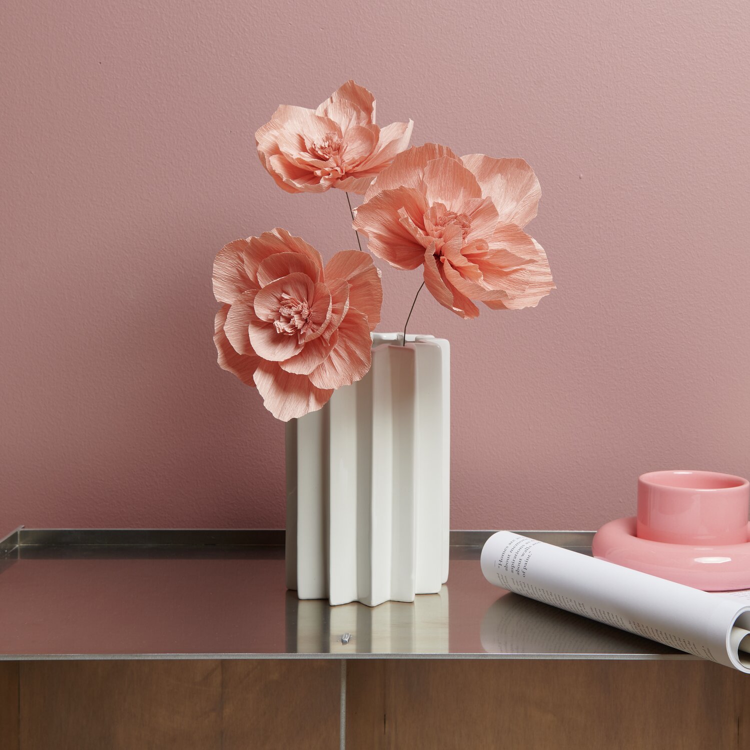 Krepppapier Blüten in Terracotta von Rico Design in einer weißen Vase auf einem Tisch vor rosa Wand.