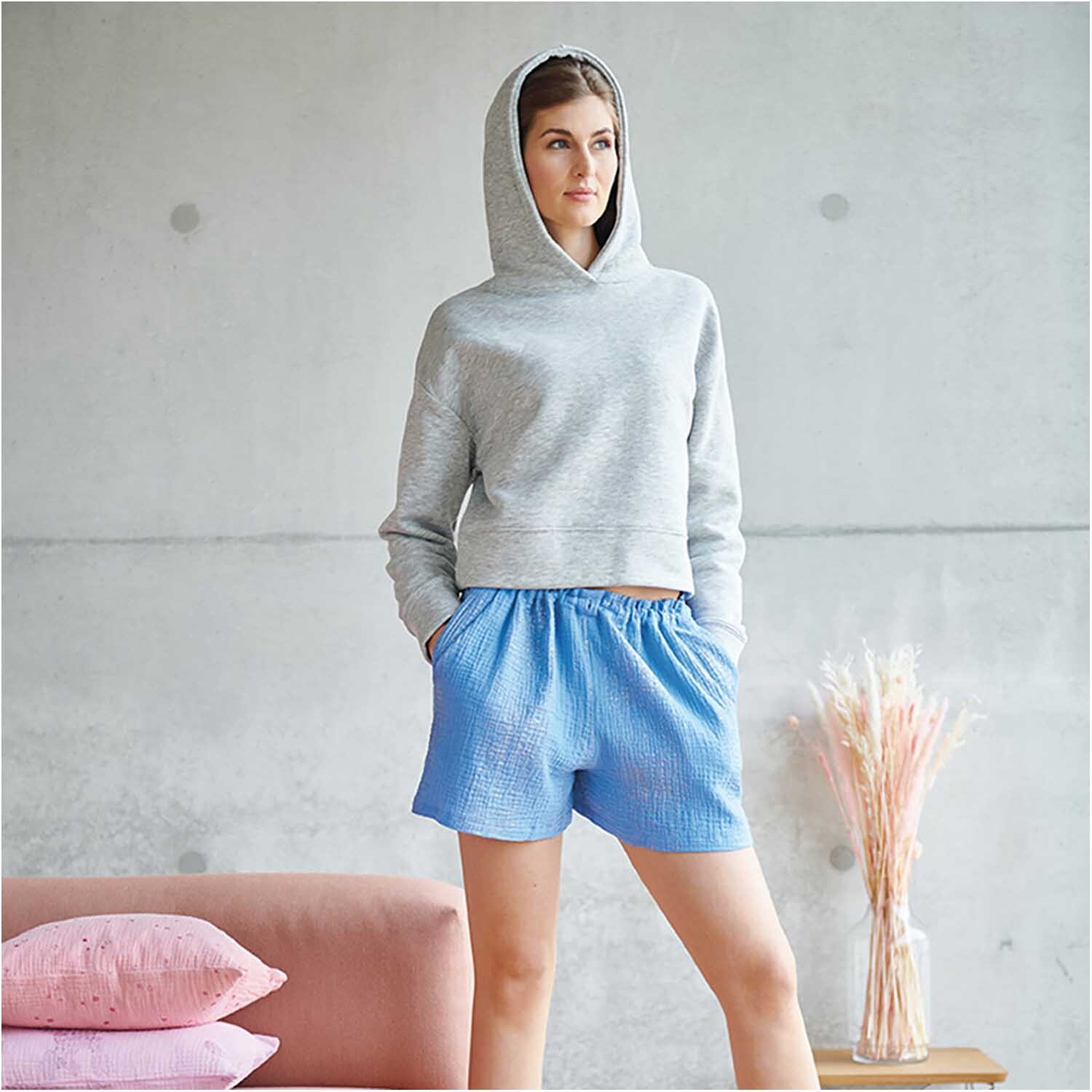 Meterware Krinkelmusselin azur Hot Foil von Rico Design Meterware Krinkelmusselin azur Hot Foil: Frau in grauem Hoodie und blauen Shorts aus Krinkelmusselin, neben rosa Kissen und Trockenblumen.