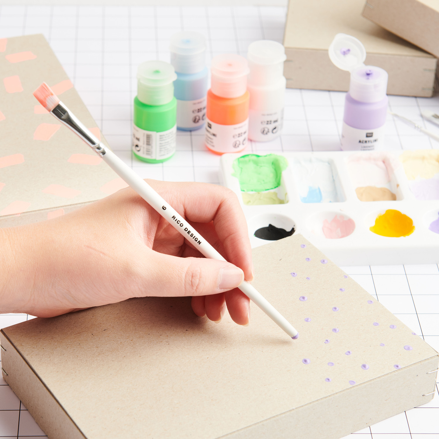 Acrylini Acrylfarben Set Power Colours 12x22ml von Rico Design Acrylini Acrylfarben Set Power Colours: Eine Hand malt mit Pinsel auf Karton, umgeben von bunten Farben und Malutensilien.