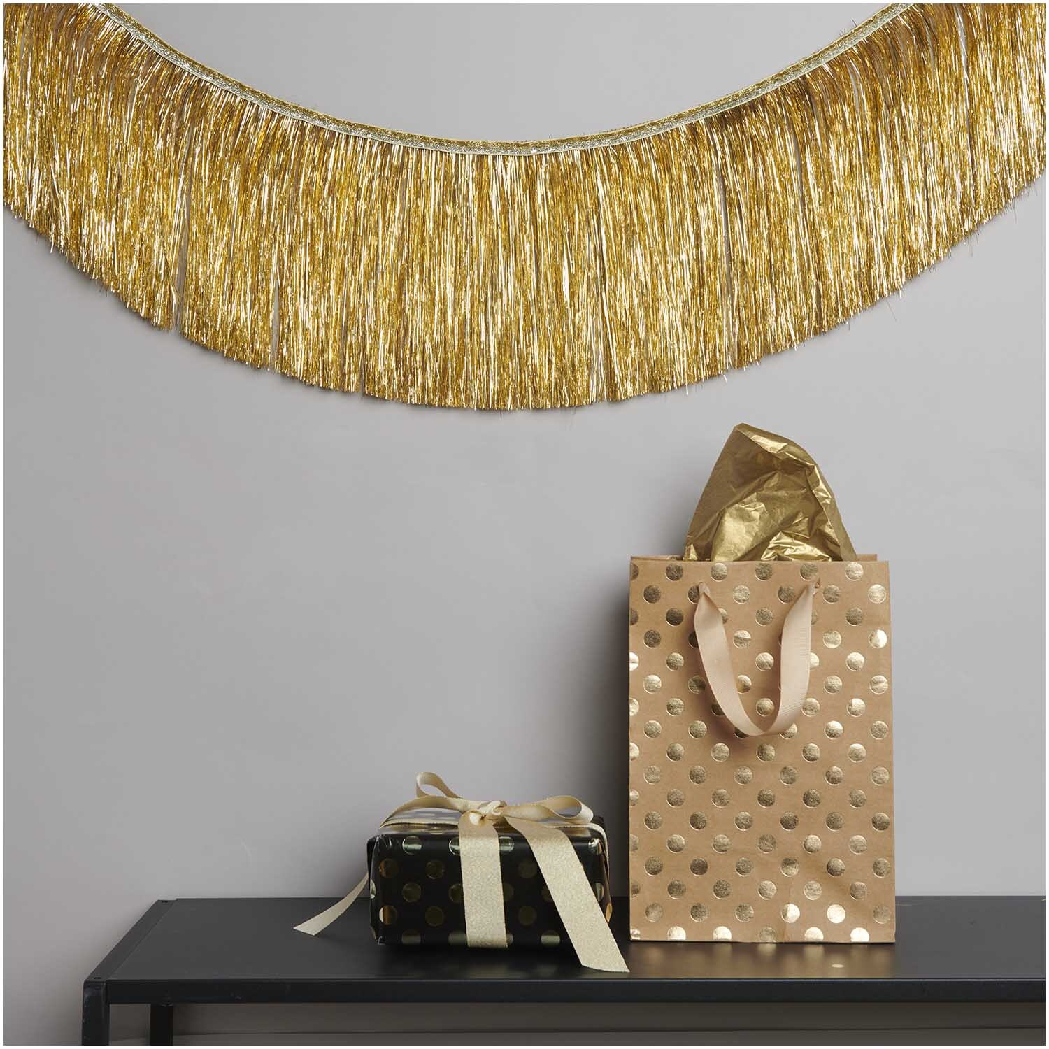 Lametta Girlande gold 3,5m von Rico Design Lametta Girlande gold 3,5m von Rico Design dekoriert eine Wand über einem Tisch mit Geschenken.