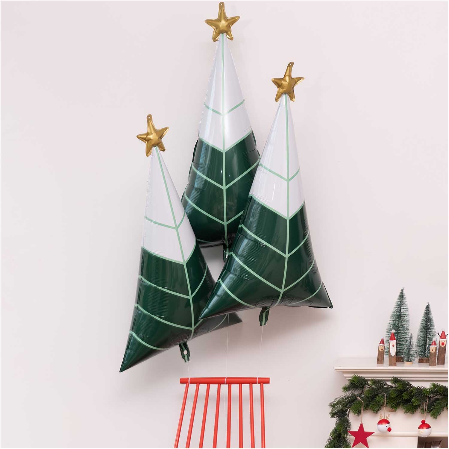 Holzhänger Rentier Kugel rot-weiß 5x7,5cm von Rico Design Holzhänger Rentier Kugel zeigt rote und weiße Details. Im Hintergrund sind grüne Weihnachtsbäume mit goldenen Sternen zu sehen.