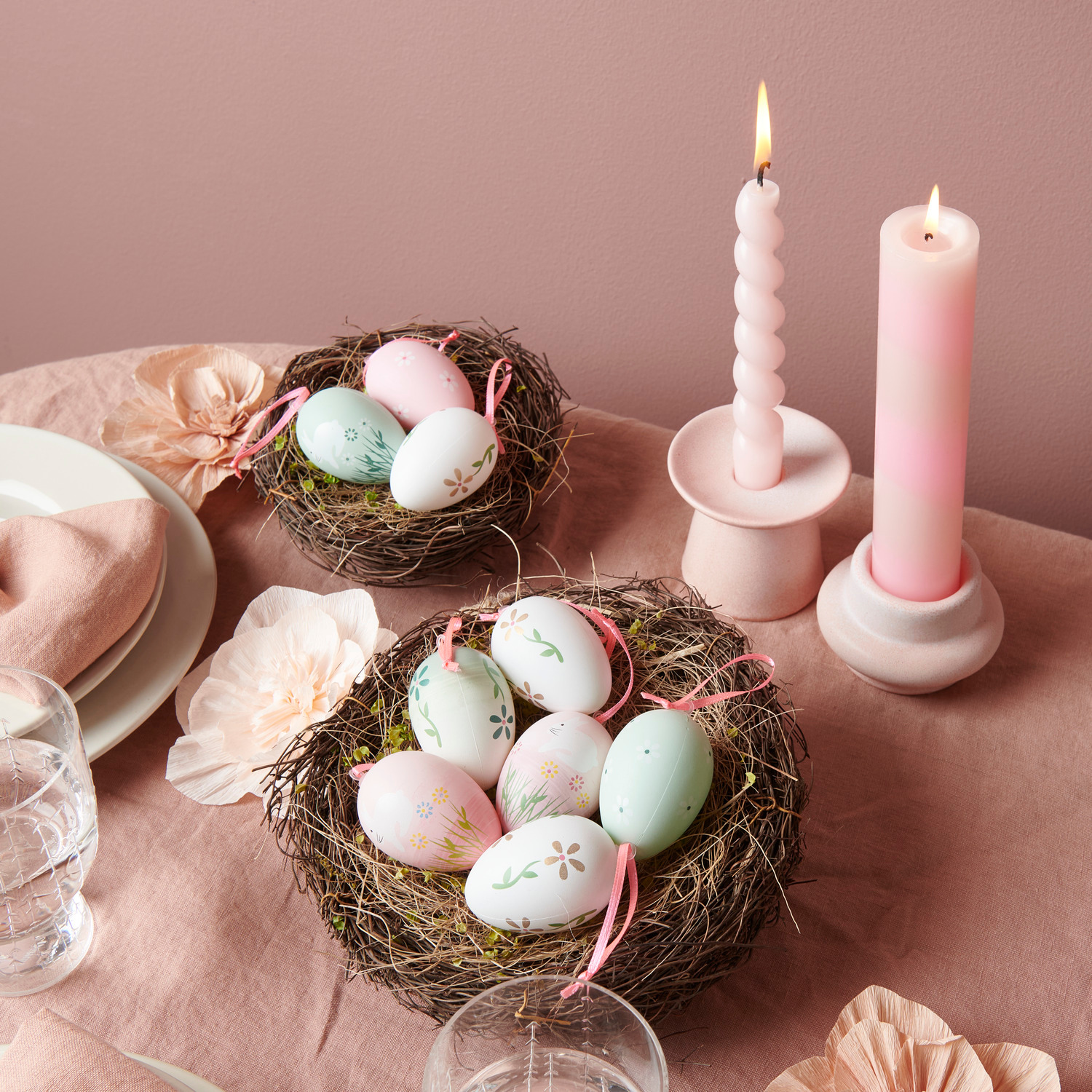 Deko-Eier Rosa in Naturnest mit Gras, umgeben von Kerzenhaltern aus Porzellan und Stabkerzen.