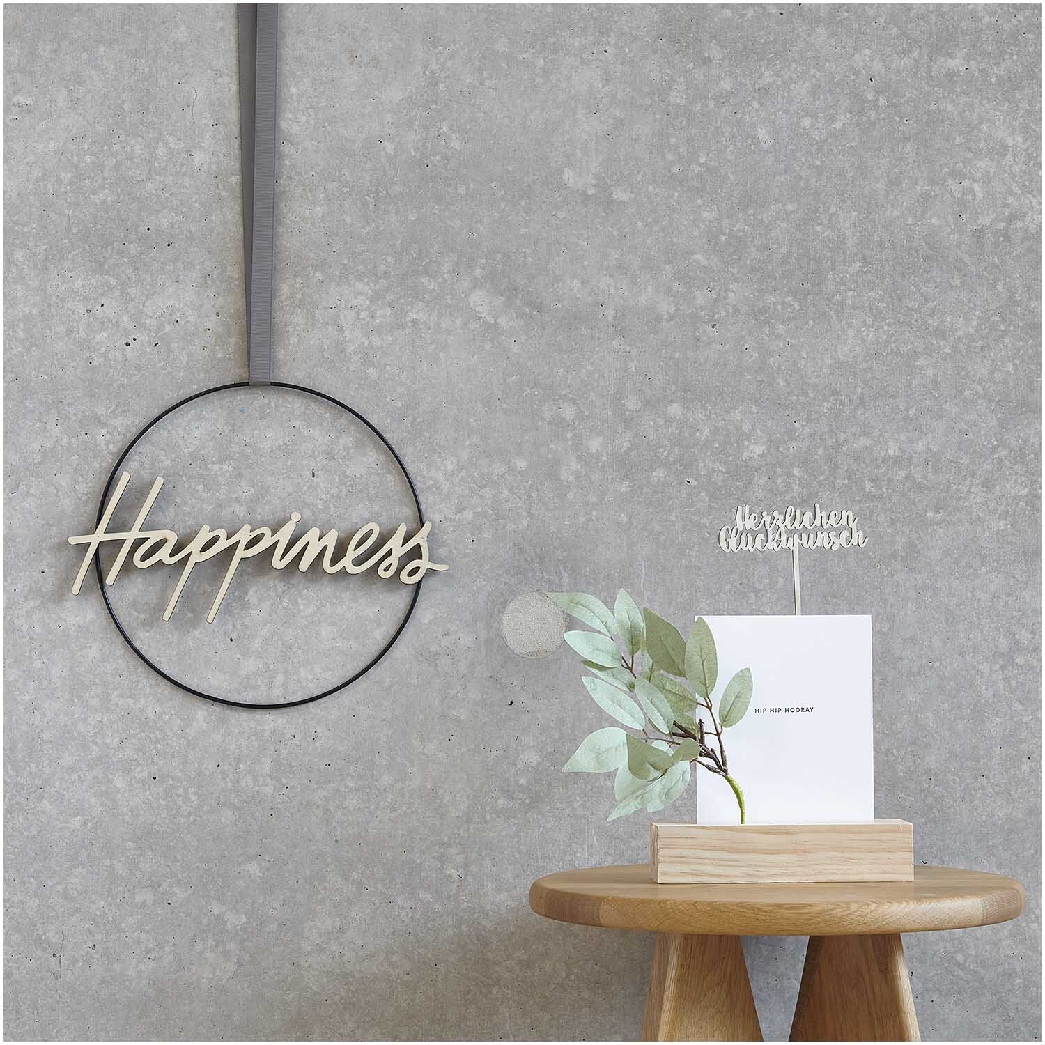 Lorbeerzweig 26cm pastellgrün von Rico Design auf einem Tisch, neben einer Karte und einem Dekoring mit "Happiness" an einer Betonwand.