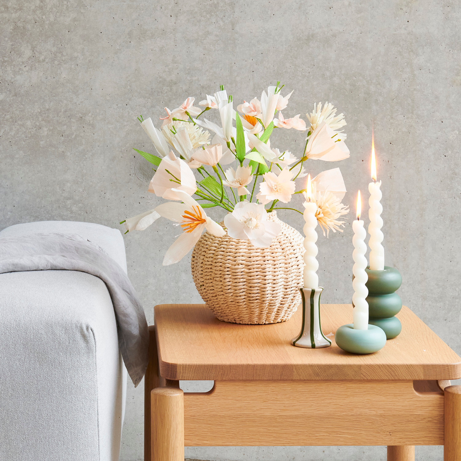 Vase Geflecht oval Natur mit Papierblumen in Creme und Weiß auf Holztisch neben grünen Porzellan Kerzenhaltern mit Spiralkerzen.