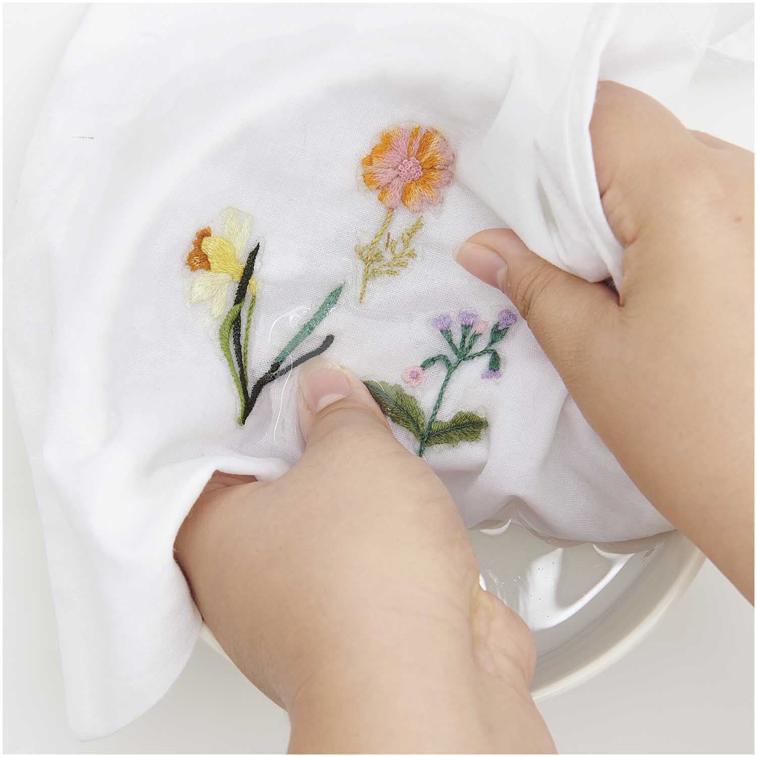 Stick and Stitch Kit de broderie Fleurs de printemps dessinées 15 pièces from Rico Design Stick and Stitch Stickpackung Frühlingsblumen zeigt bunte Blumenstickerei auf weißem Stoff. Hände halten das Tuch, ideal für kreative Projekte.