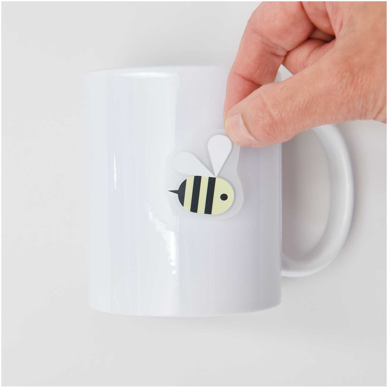 Transfermotiv Biene 15 Stück von Rico Design Transfermotiv Biene: Eine Hand klebt ein Bienenmotiv auf eine weiße Tasse. Marke: Rico Design.