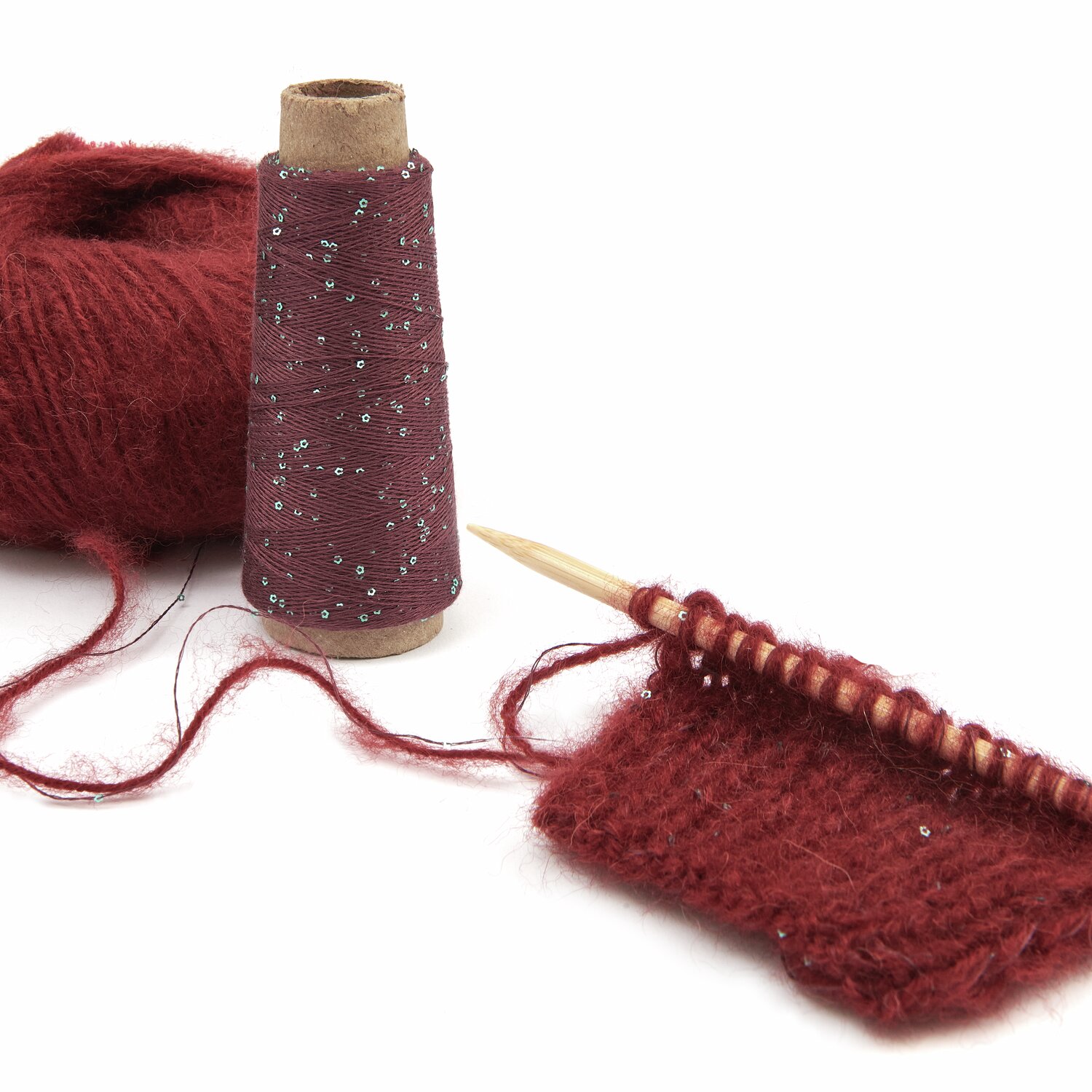 Luxury Merino Alpaca Dream aran 50g 125m Farbe Bordeaux von Rico Design Rotes Strickgarn mit Glitzerfaden, auf einer Holzstricknadel gestrickt. Daneben eine Garnrolle. Perfekt für kreative Strickprojekte. Marke: Unbekannt.