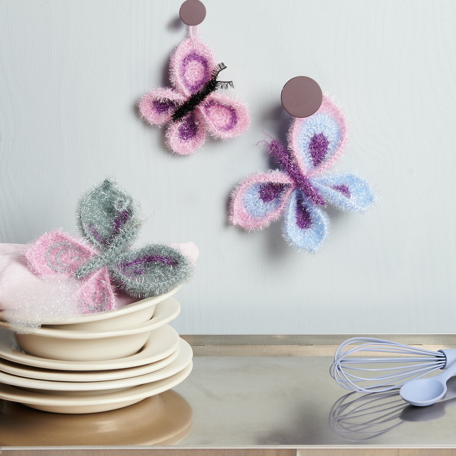 Häkelset Creative Bubble Schmetterling von Rico Design Häkelset Creative Bubble Schmetterling von Rico Design: Zwei gehäkelte Schmetterlinge in Pastellfarben, dekorativ an der Wand und auf Geschirr platziert.