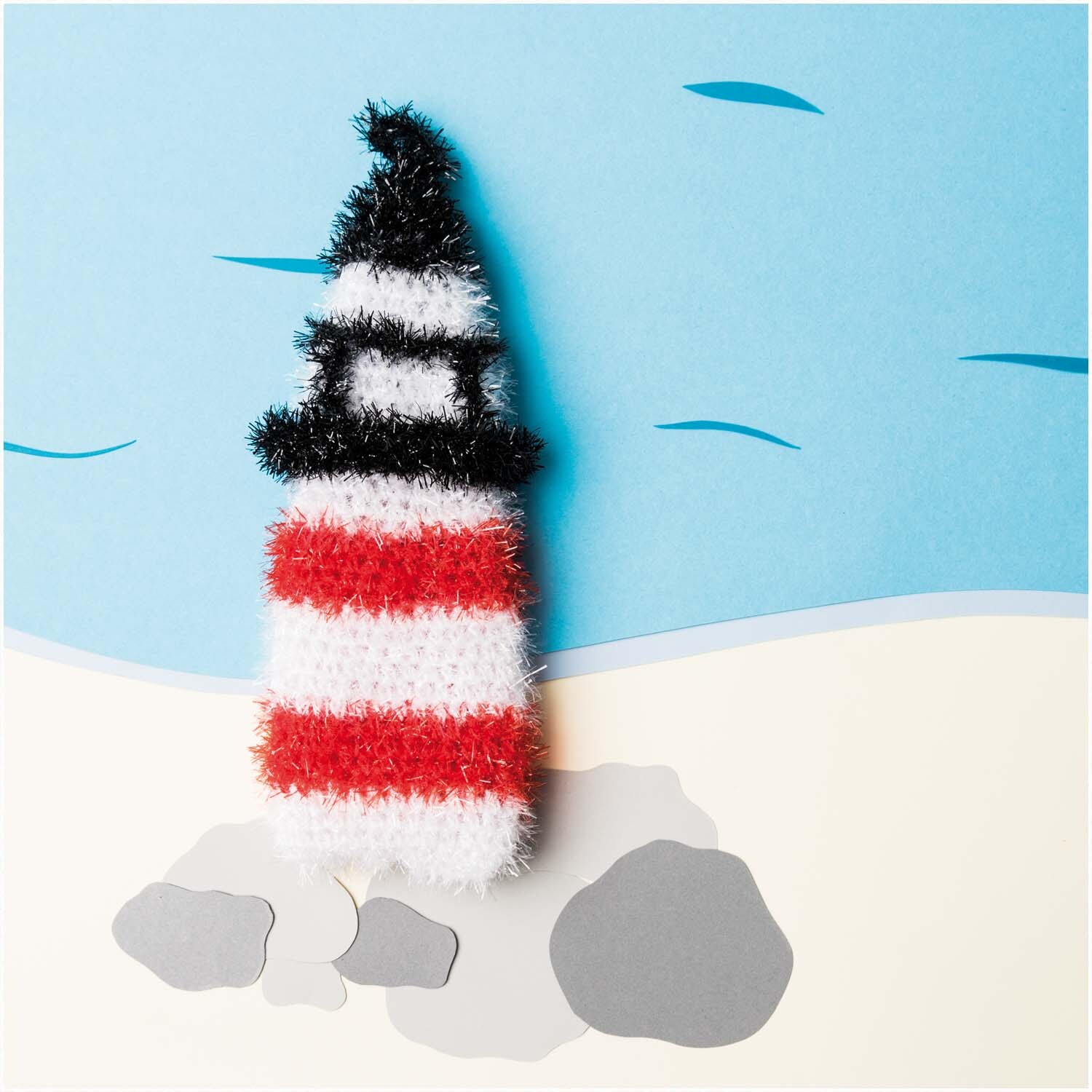 Creative Bubble - At the Beach: Ein gestrickter Leuchtturm in Rot, Weiß und Schwarz von Rico Design am Strand.