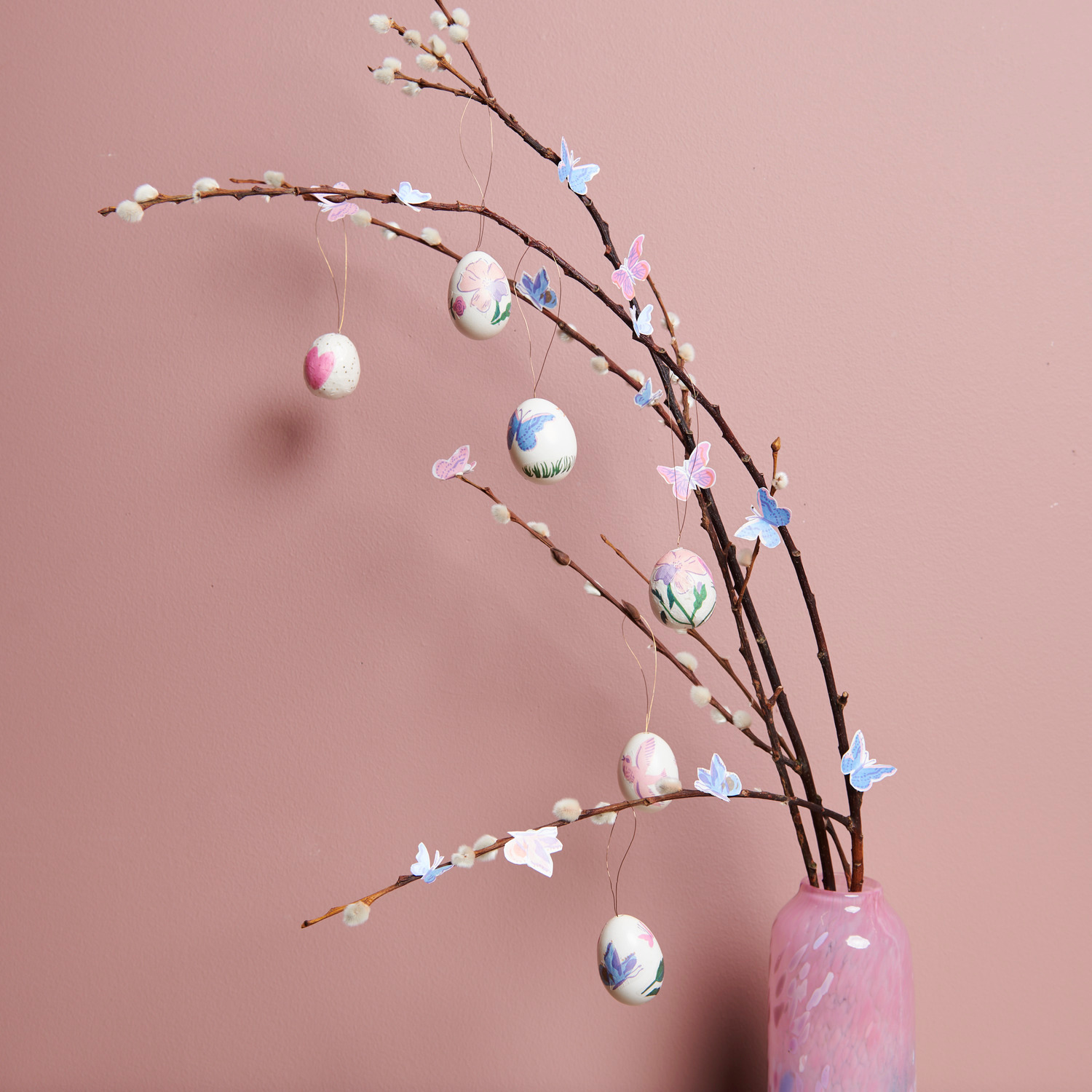 Paper Patch Papier Frühlingswiese Kerzenwachs Granulat: Dekorative Zweige mit bemalten Eiern und Schmetterlingen in einer rosa Vase.