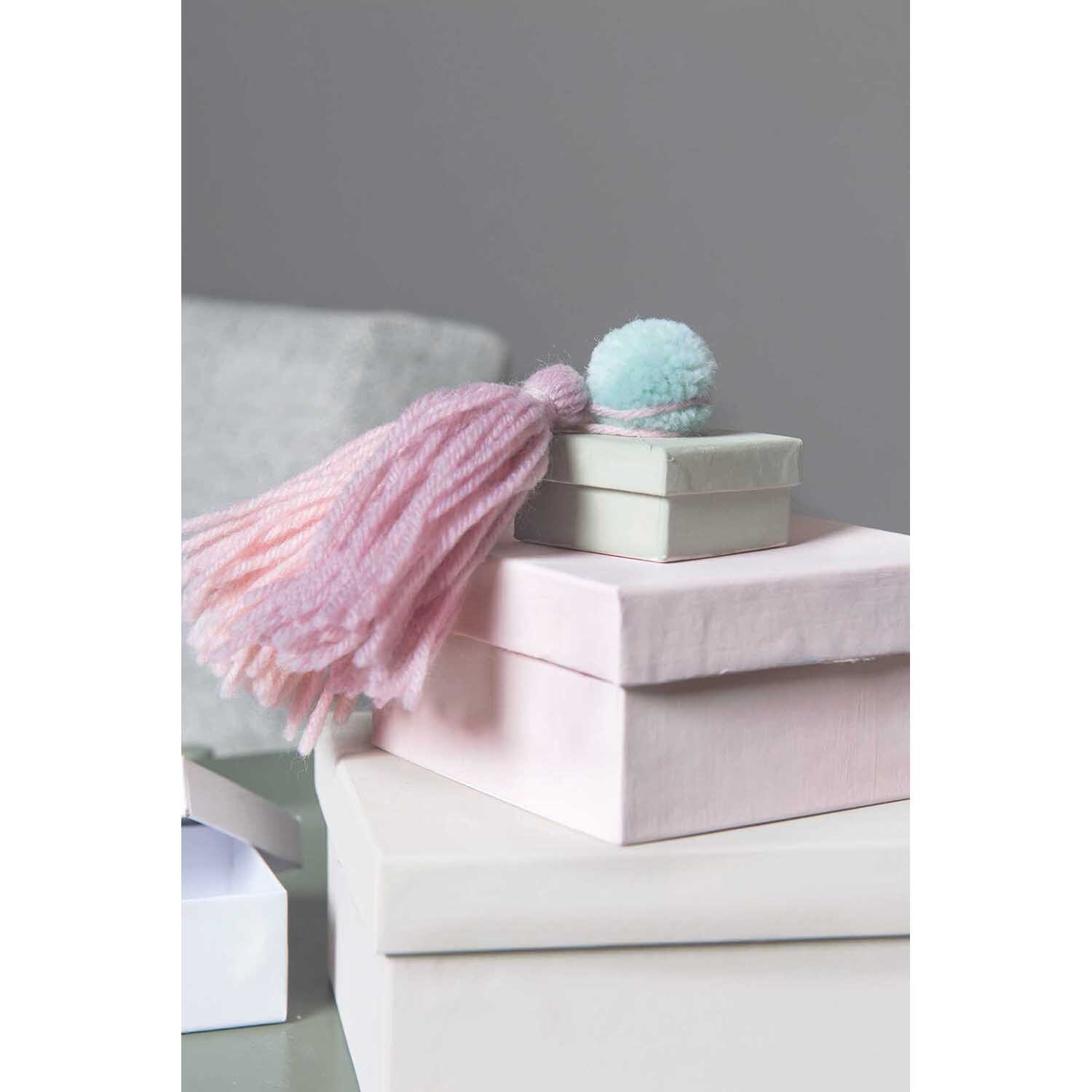 Garn Pompons 24 Stück Farbe Natur Mix von Rico Design Garn Pompons 24 Stück von Rico Design auf gestapelten Geschenkboxen mit rosa Quaste und blauem Pompon.