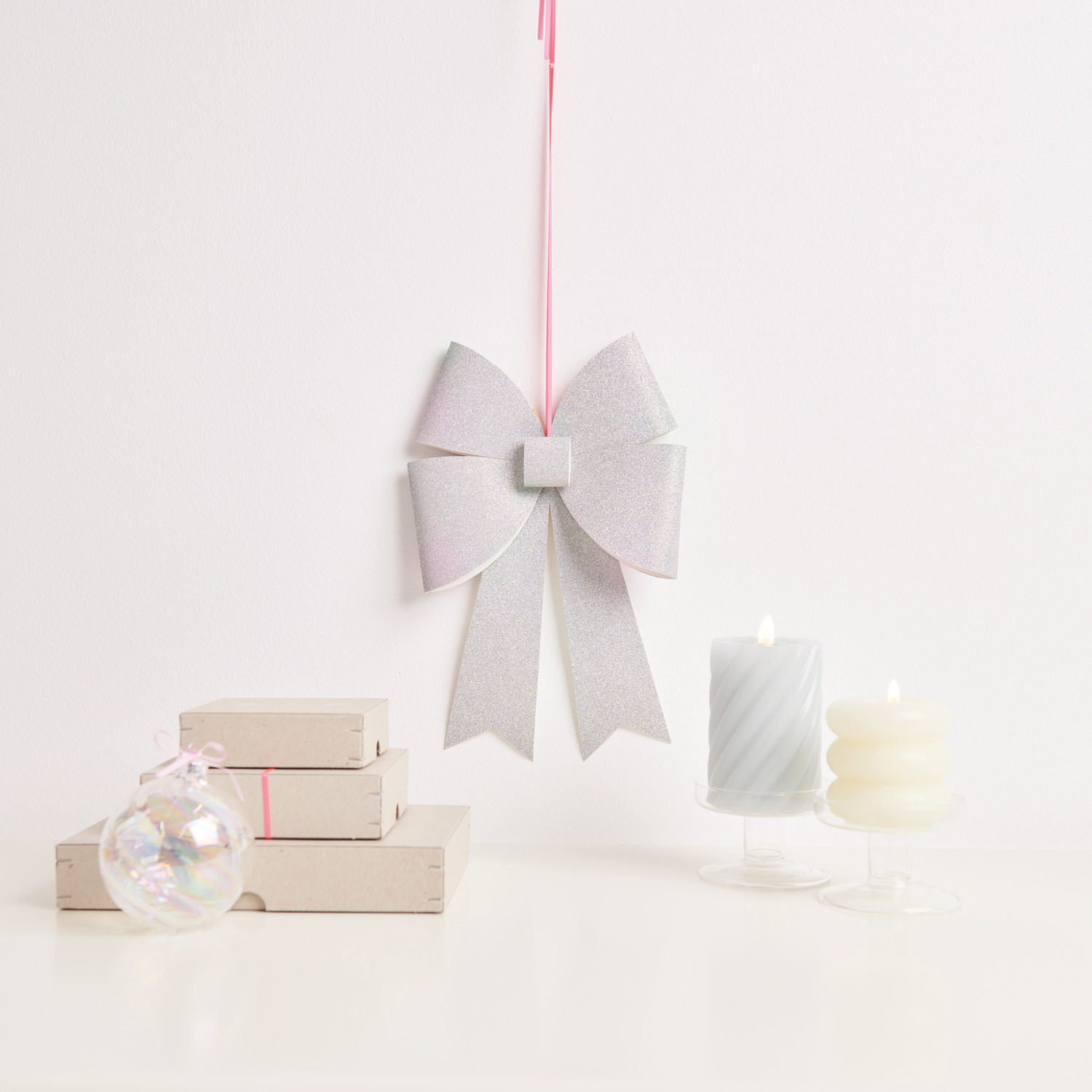 Paper Poetry Bastelset XL-Schleife Glitter 20x30cm Farbe Silber Irisierend von Rico Design Paper Poetry Bastelset XL-Schleife Glitter, silber irisierend, hängt neben Kerzen und Geschenken.