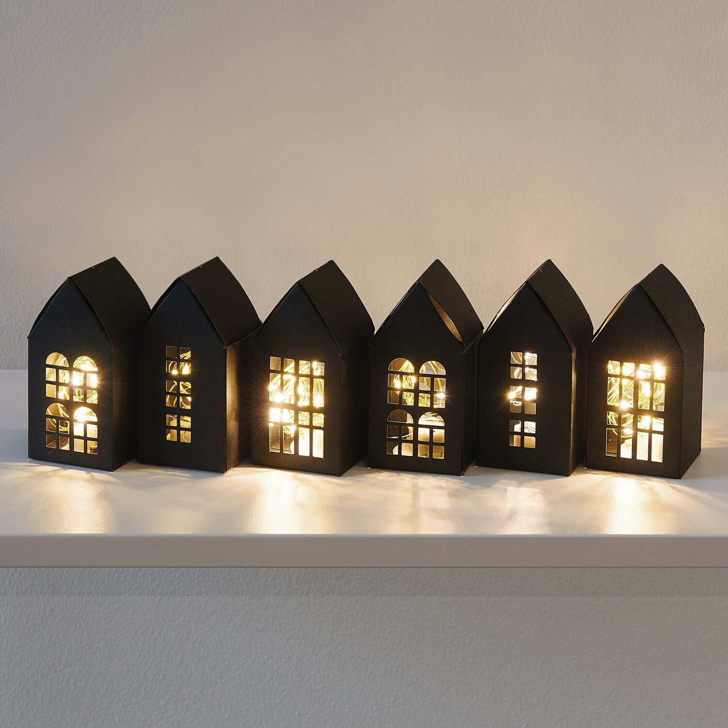 Paper Poetry Papphäuser-Set vorgestanzt 9x4,5x4,5cm 6 Stück Farbe Schwarz von Rico Design Paper Poetry Papphäuser-Set: Sechs schwarze, beleuchtete Papphäuser auf einem Regal, ideal als Adventskalender.