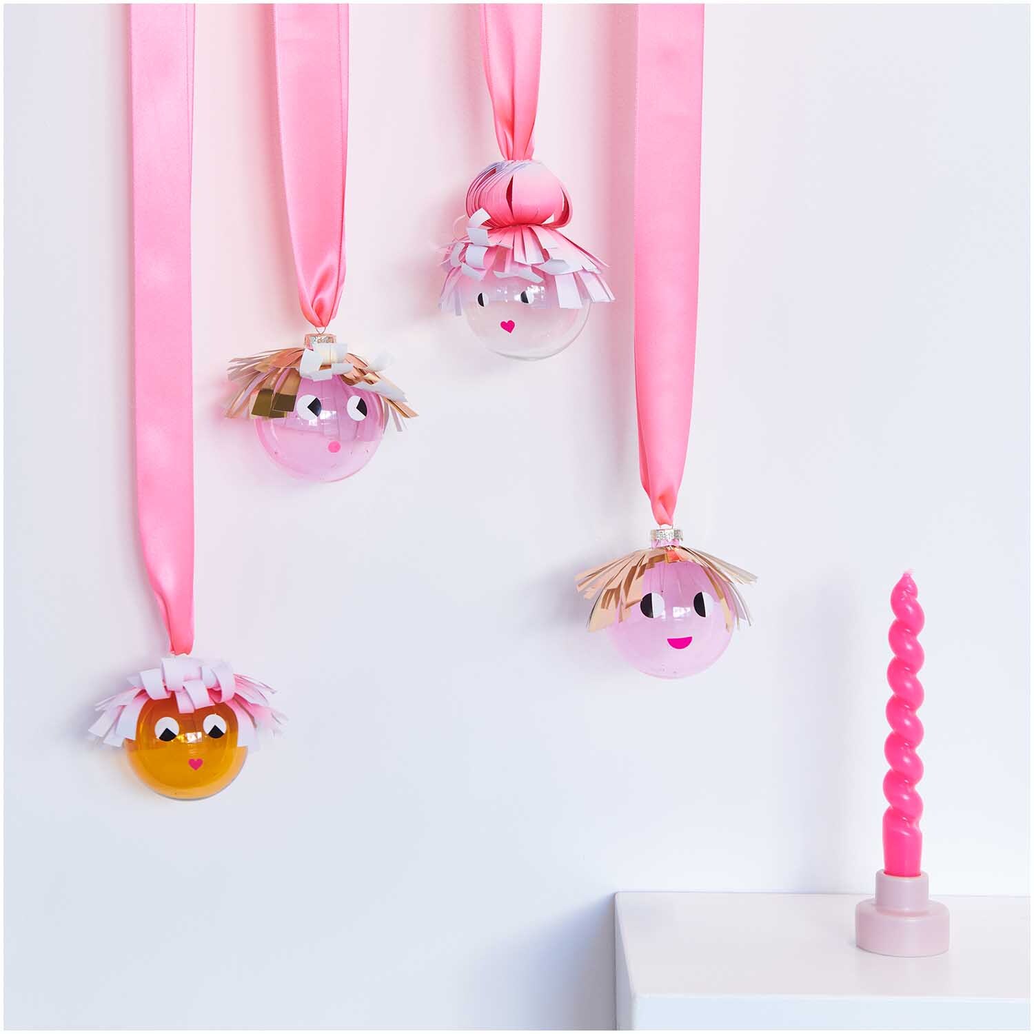 Glaskugel Baumschmuck einfarbig 8cm Farbe Puder Transparent von Rico Design Glaskugel Baumschmuck einfarbig 8cm: Vier dekorative Kugeln mit Gesichtern, aufgehängt an rosa Bändern, neben einer rosa Kerze.