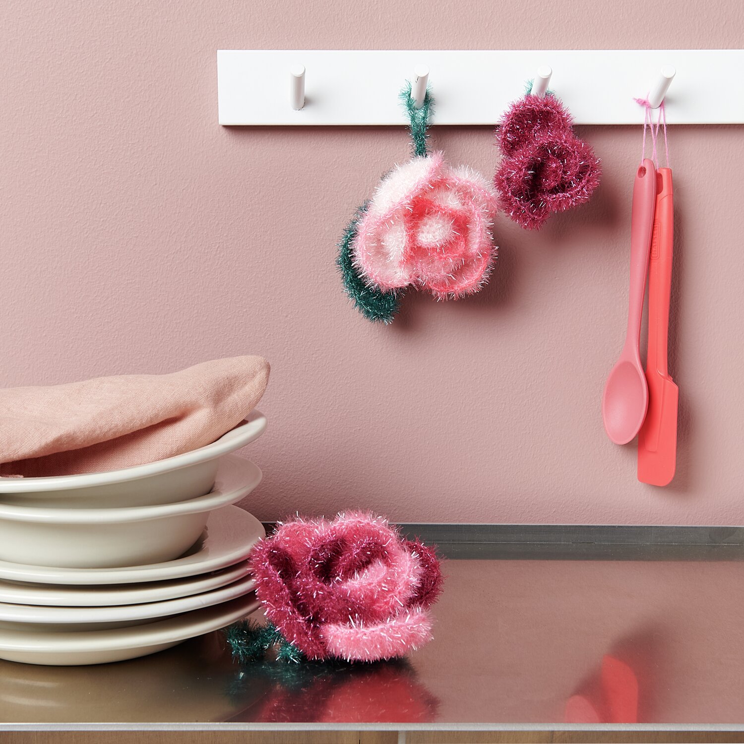 Häkelset Creative Bubble Rose Classic von Rico Design Häkelset Creative Bubble Rose Classic von Rico Design: Rosa und rote gehäkelte Rosen an Haken, mit Geschirr und Küchenutensilien auf Metalloberfläche.