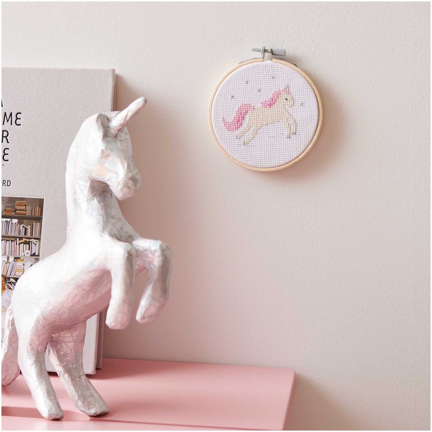 Mini Kit de broderie image licorne Ø 10cm from Rico Design Stickpackung Mini Kit Einhorn Bild Ø 10cm an einer Wand, daneben eine weiße Einhornfigur.