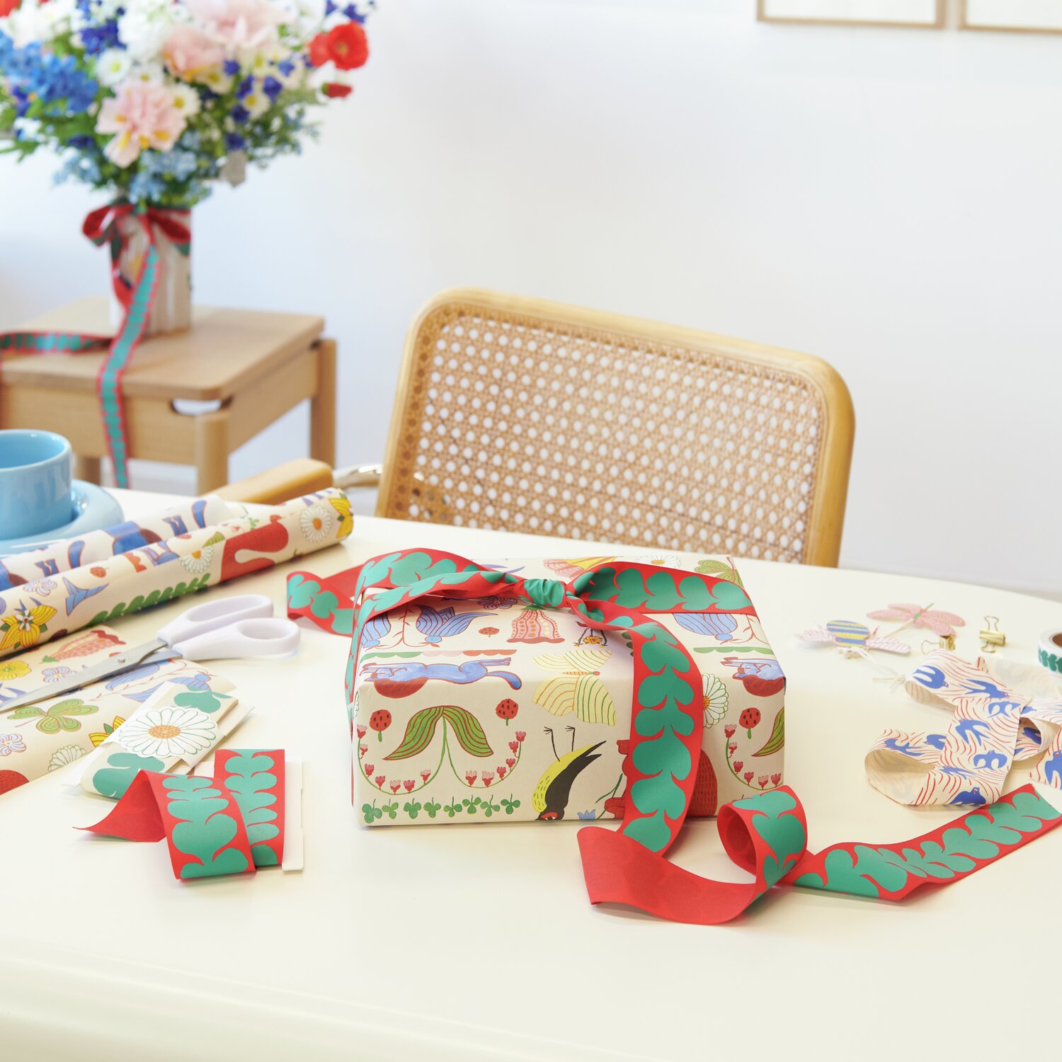 Geschenkpapier Midsommar Magic 70cm 2m 100g/m² von Rico Design Geschenkpapier Midsommar Magic von Rico Design auf einem Tisch mit Geschenkverpackung und bunten Bändern. Im Hintergrund ein Blumenstrauß.