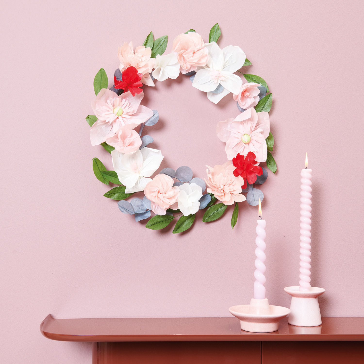 Blütenkranz Papier Rosé, dekorativ an der Wand, mit Porzellankerzenhalter und Spiralkerzen in Rosa auf einem Tisch arrangiert.