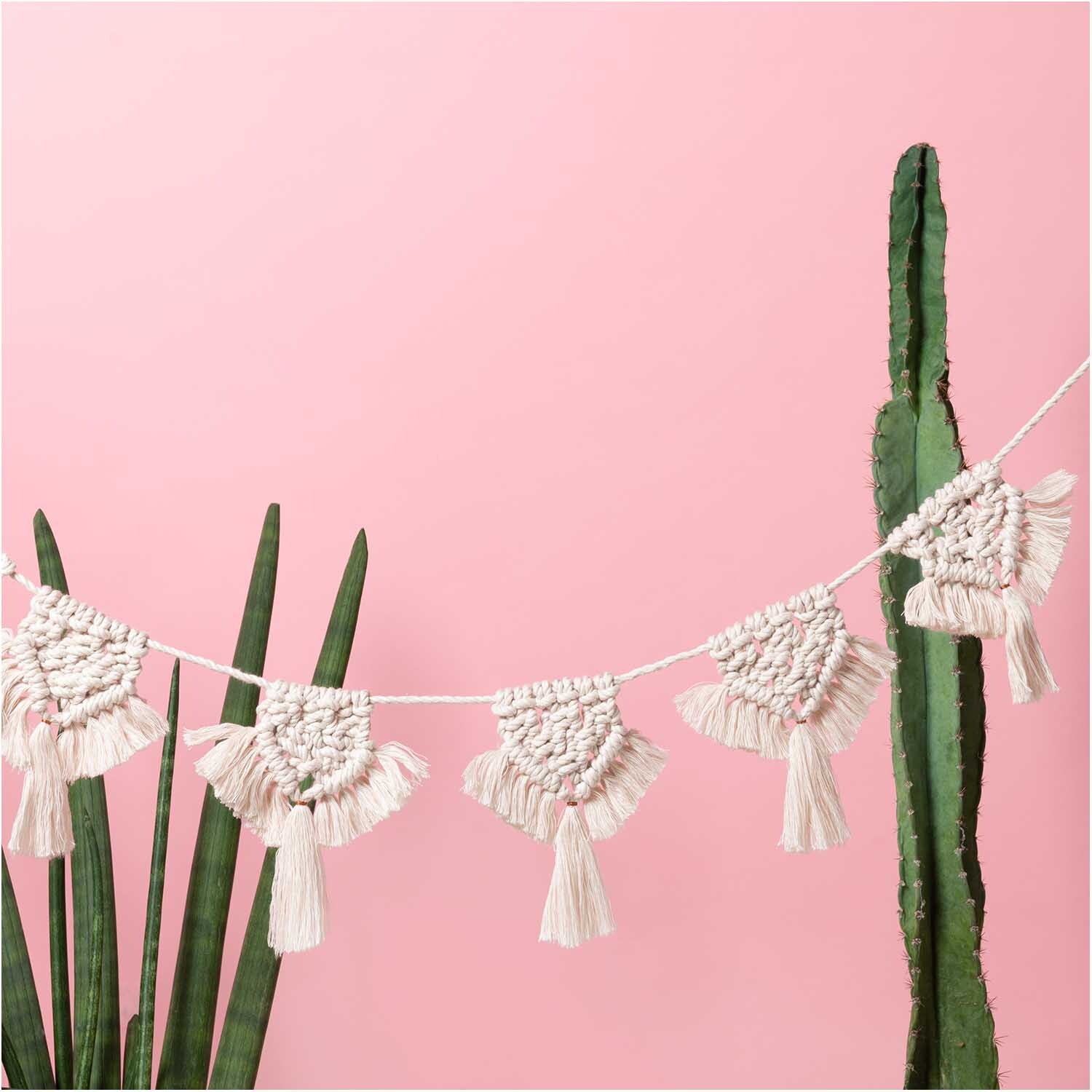 Creative Cotton Cord Makramee-Garn von Rico Design, dekorativ als Girlande vor rosa Hintergrund mit Pflanzen.