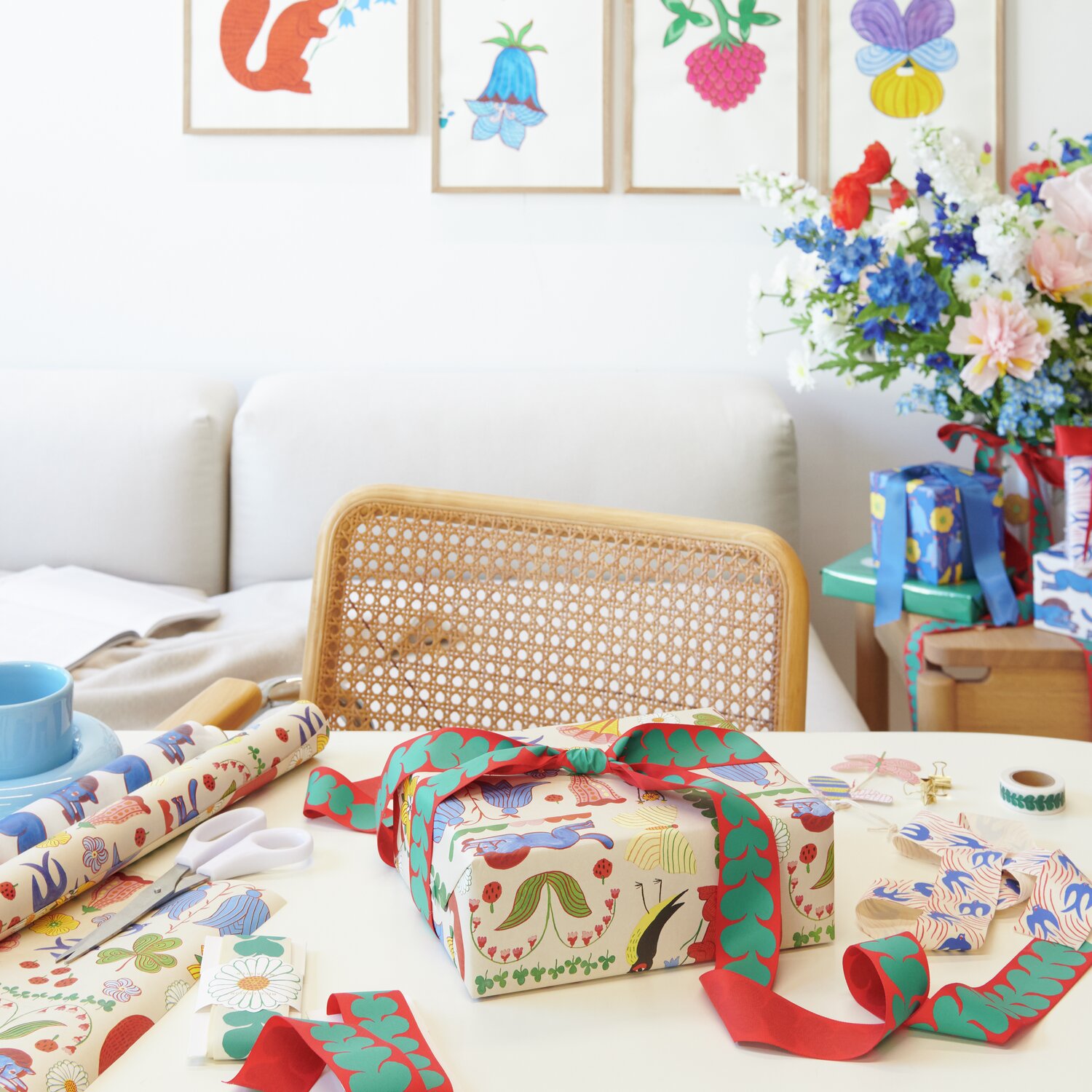 Geschenkpapier Midsommar Magic 70cm 2m 100g/m² von Rico Design Geschenkpapier Midsommar Magic auf einem Tisch mit bunten Blumenmustern, umgeben von Bastelmaterialien und einem Geschenk.