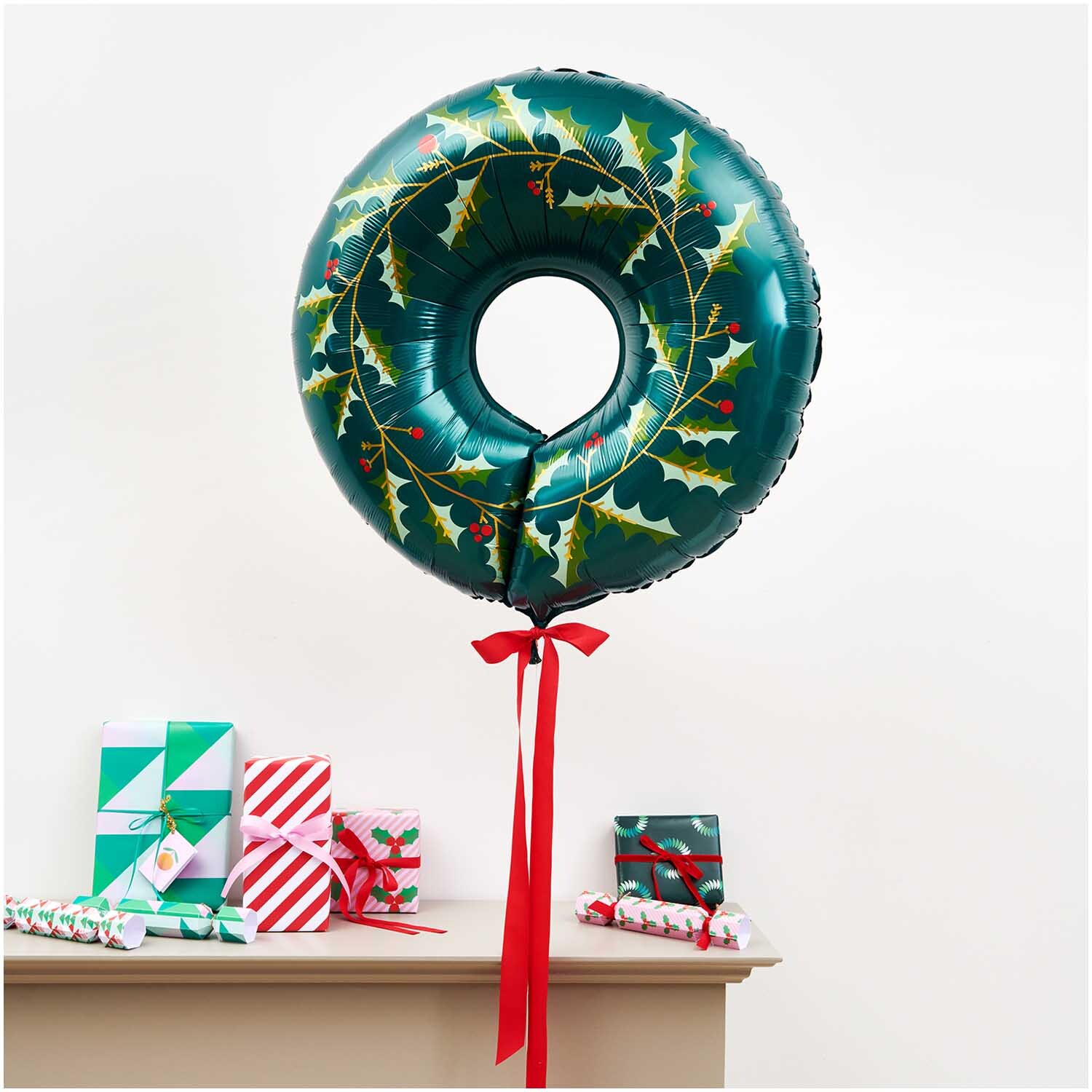 Folienballon Weihnachten Kranz 71x20cm von Rico Design Folienballon Weihnachten Kranz 71x20cm in Grün mit Stechpalmenmotiv, dekoriert mit roten Bändern, schwebt über Geschenken auf einem Tisch.