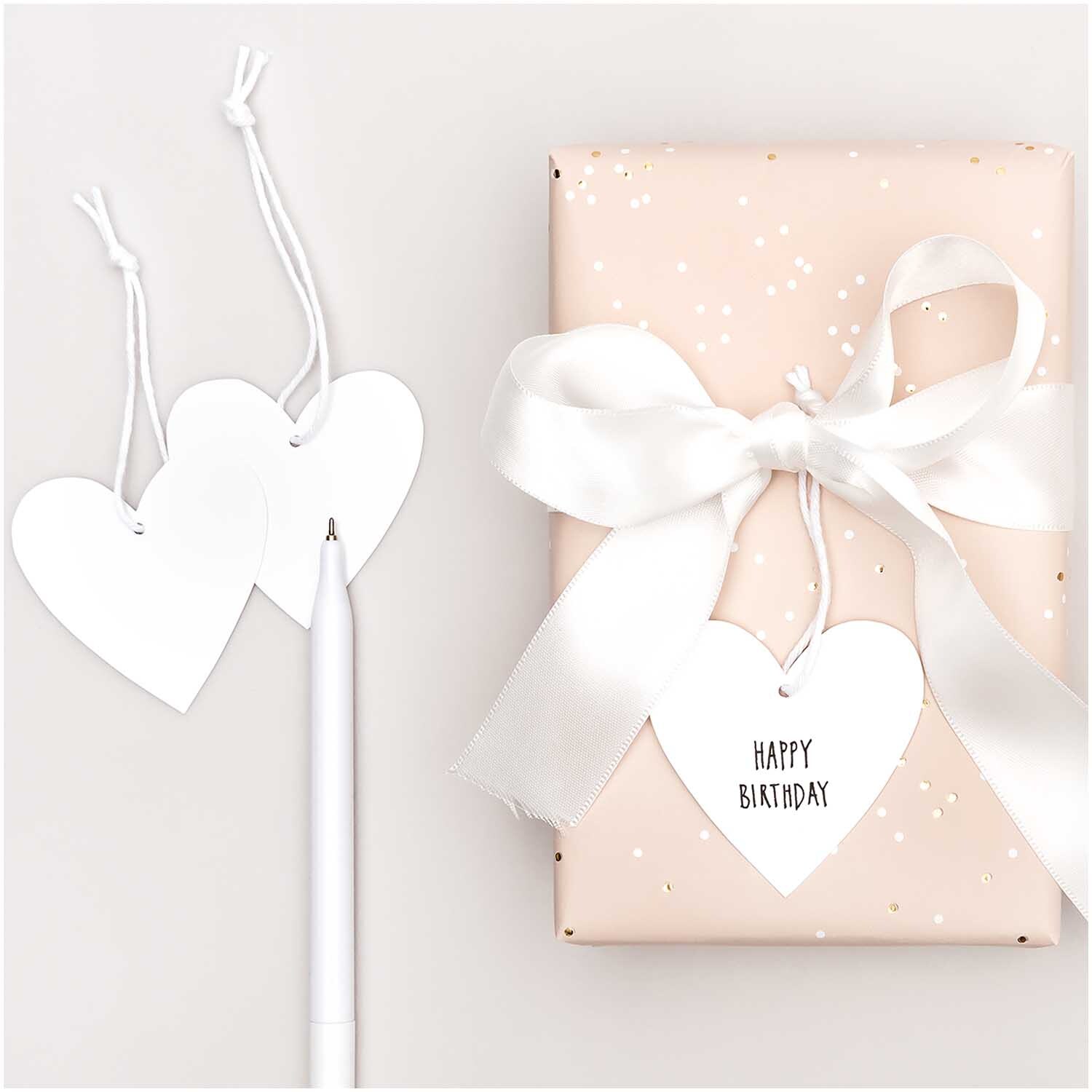 Paper Poetry Papieranhänger Herzen M 24 Stück Farbe Weiß von Rico Design Paper Poetry Papieranhänger Herzen M 24 Stück: Weiße Herzanhänger neben Geschenk mit Schleife. Ideal für kreative Dekorationen.