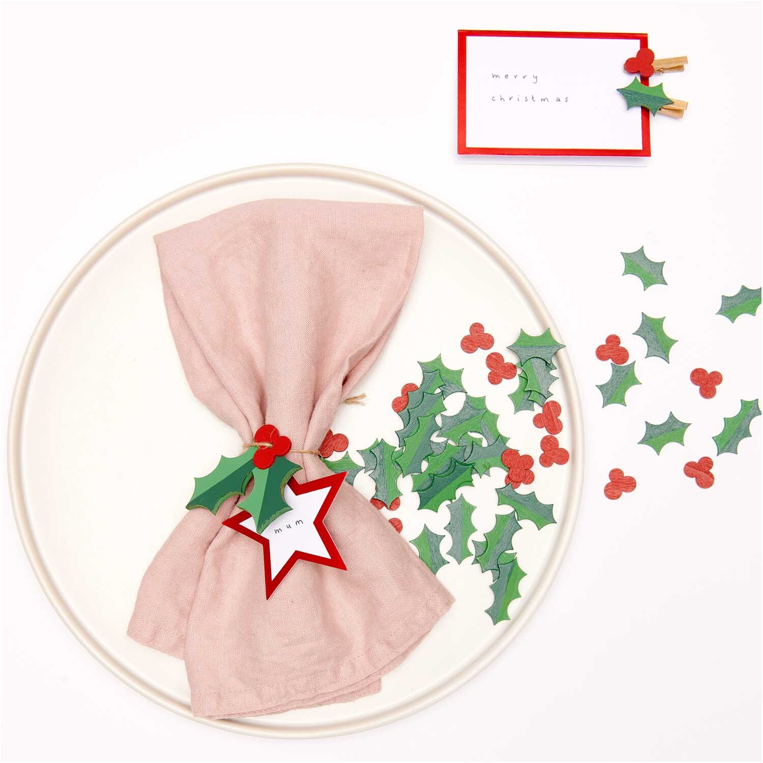 Holzhänger Ilex grün-rot 3 Stück von Rico Design Holzhänger Ilex grün-rot 3 Stück von Rico Design, dekoriert mit grünen und roten Ilexblättern auf einem Tisch mit rosa Serviette.