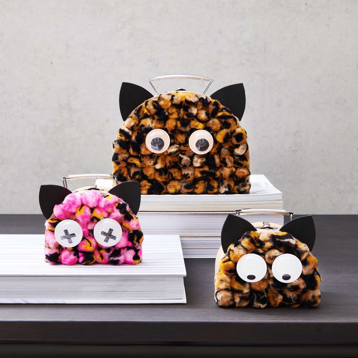 Pompons Acid Leo 25mm 40 Stück Farbe Braun-Schwarz von Rico Design Pompons Acid Leo 25mm 40 Stück: Drei flauschige Taschen mit Tierohren und Augen auf gestapelten Büchern.