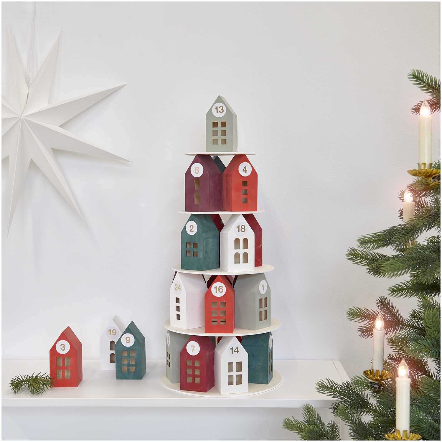 Adventskalender Holz-Haus-Pyramide von Rico Design Adventskalender Holz-Haus-Pyramide mit bunten Häuschen, dekoriert neben einem Weihnachtsbaum und Kerzen.