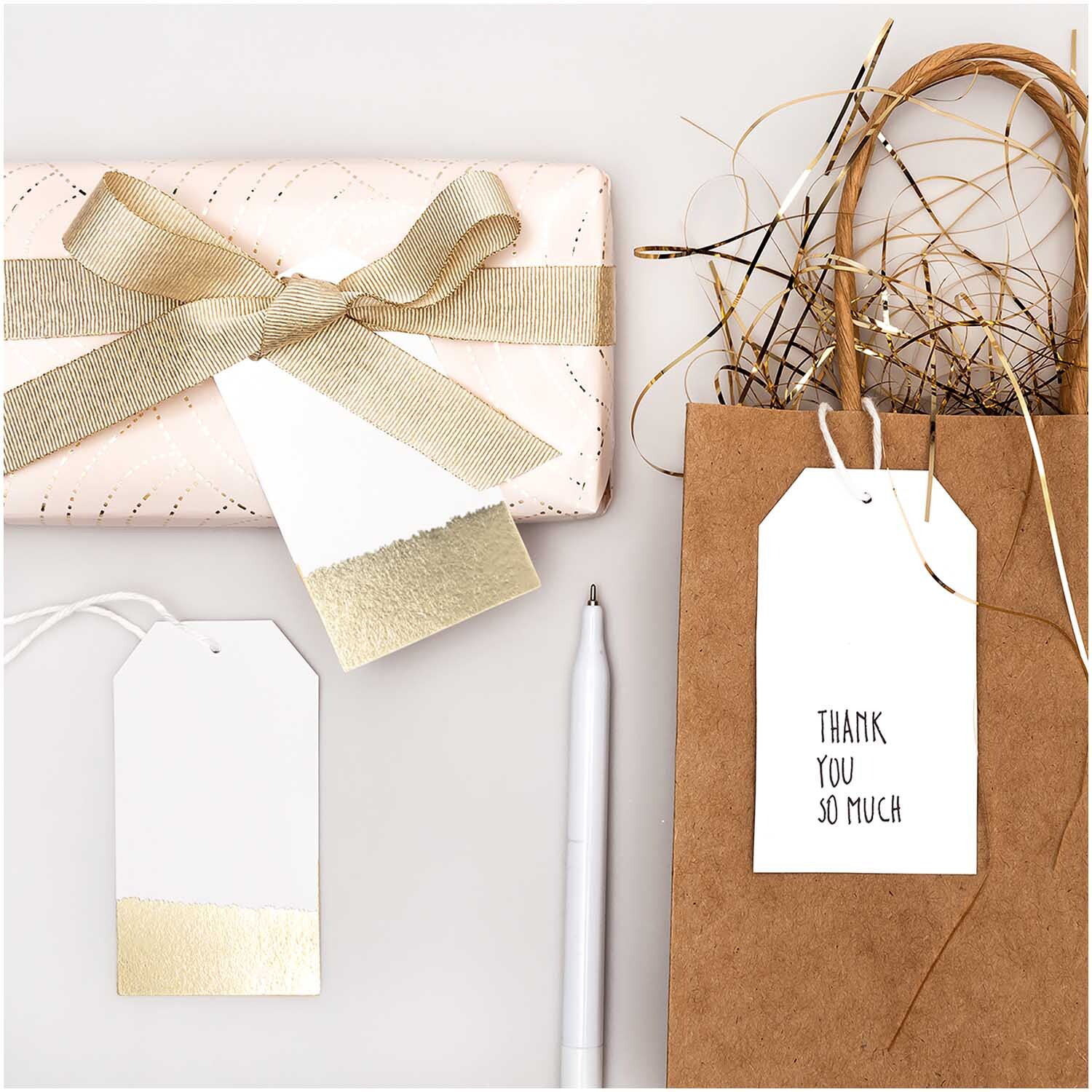 Paper Poetry Papieranhänger 4,5x8cm 24 Stück Farbe Weiß-Gold von Rico Design Paper Poetry Papieranhänger 4,5x8cm 24 Stück in Weiß-Gold neben Geschenk und Tüte. Ideal für kreative Verpackungen.
