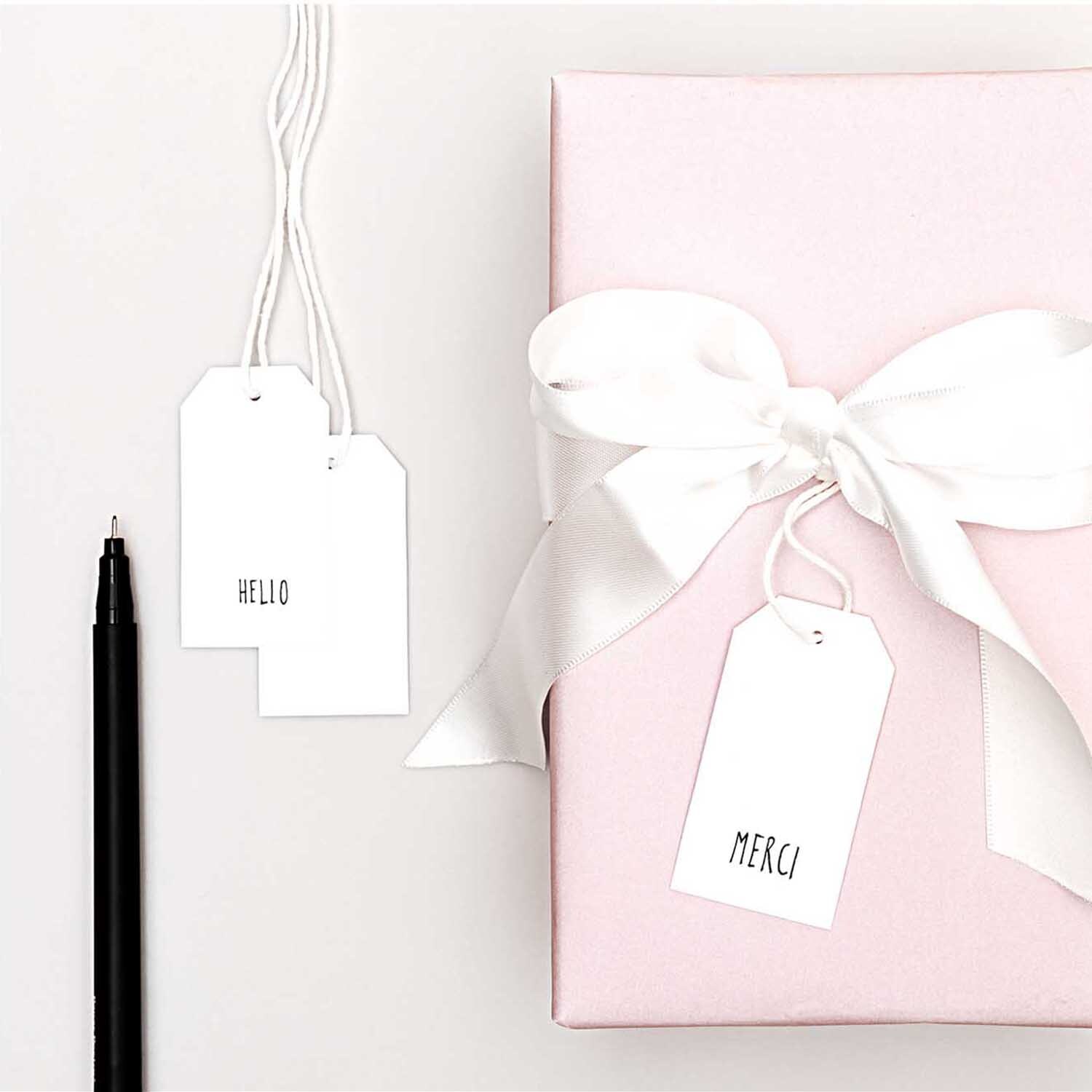 Paper Poetry Papieranhänger weiß 24 Stück Größe 3,5x6cm von Rico Design Paper Poetry Papieranhänger weiß 24 Stück neben rosa Geschenk mit weißer Schleife und schwarzem Stift.