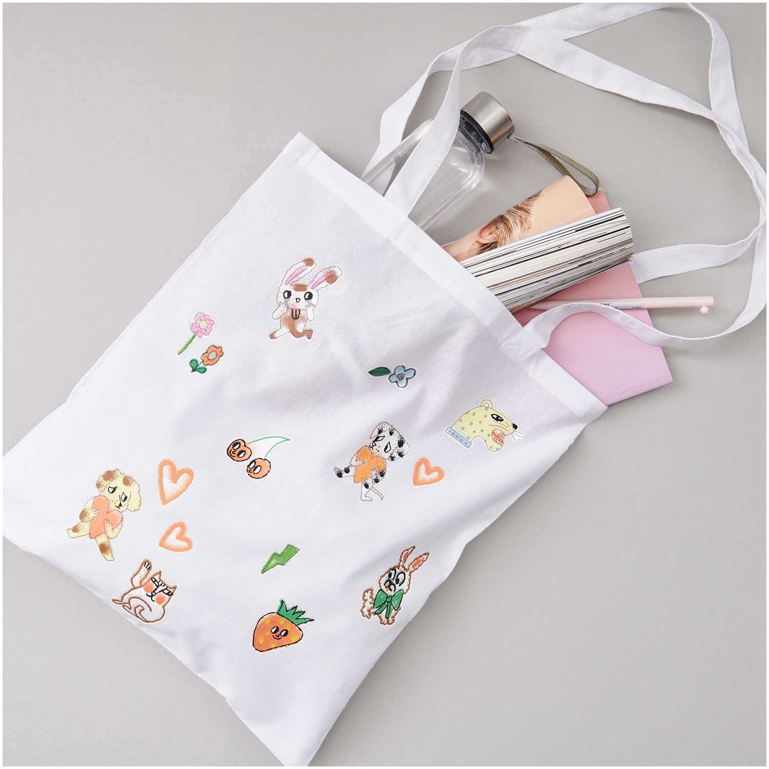 Bügelmotiv Airbrush 15 Stück: Weiße Stofftasche mit bunten Tier- und Herzmotiven, gefüllt mit Büchern und einer Wasserflasche.
