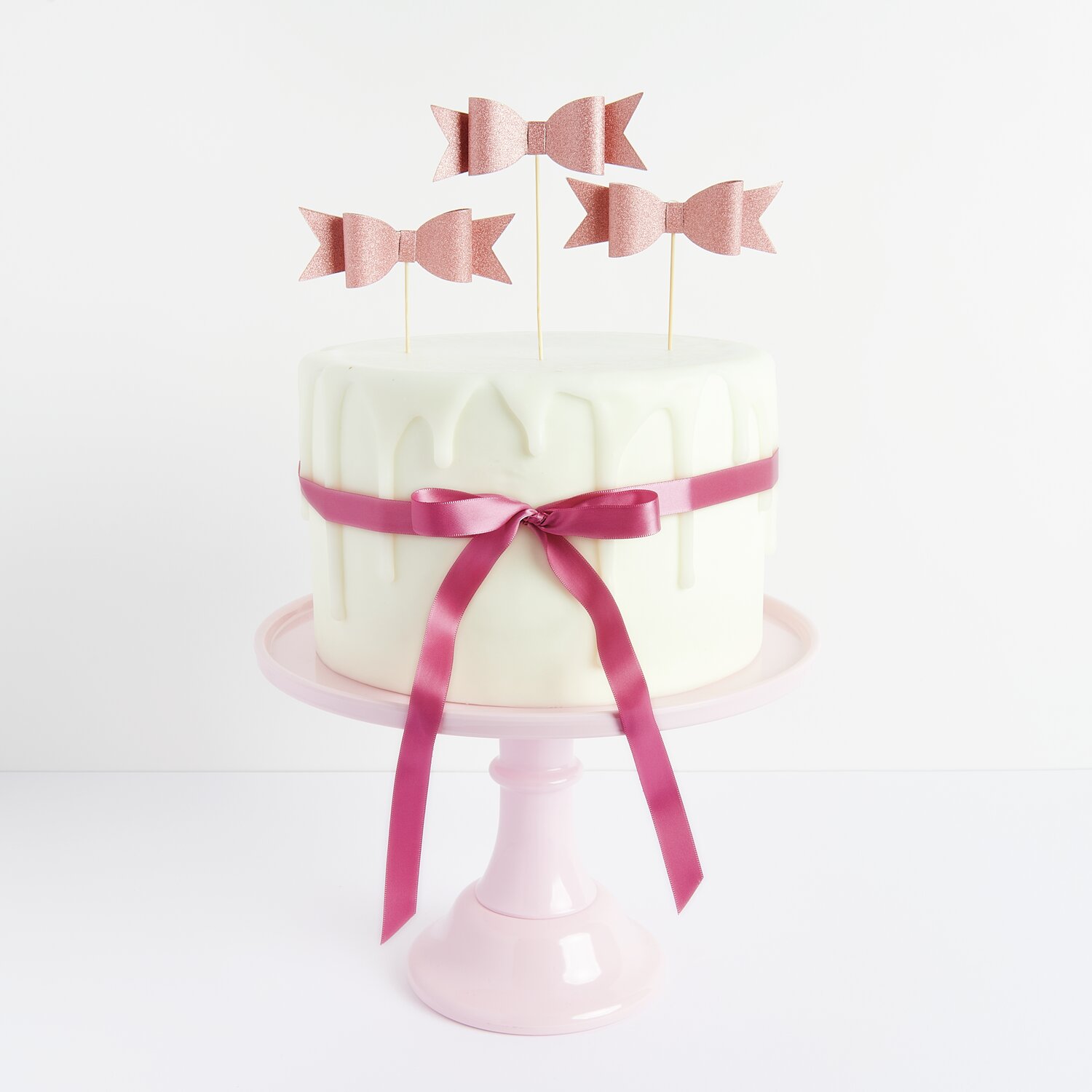 Kuchenkerzen Rosen 5 Stück von Rico Design Kuchenkerzen Rosen auf einem weißen Kuchen mit roségoldener Schleife, präsentiert auf einem rosa Tortenständer.
