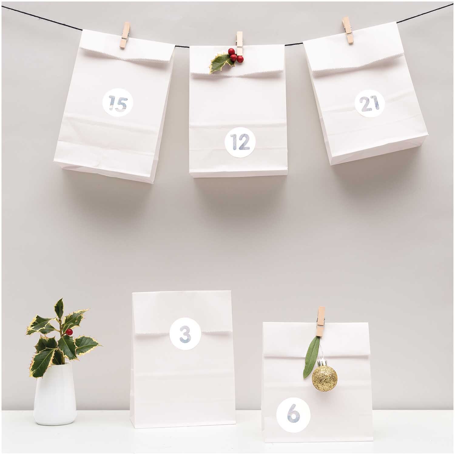 Paper Poetry Adventskalenderset weiß 24 Tüten inkl. Zubehör: Weiße Tüten mit Zahlen, dekoriert mit Klammern und Zweigen, auf einer Leine aufgehängt.