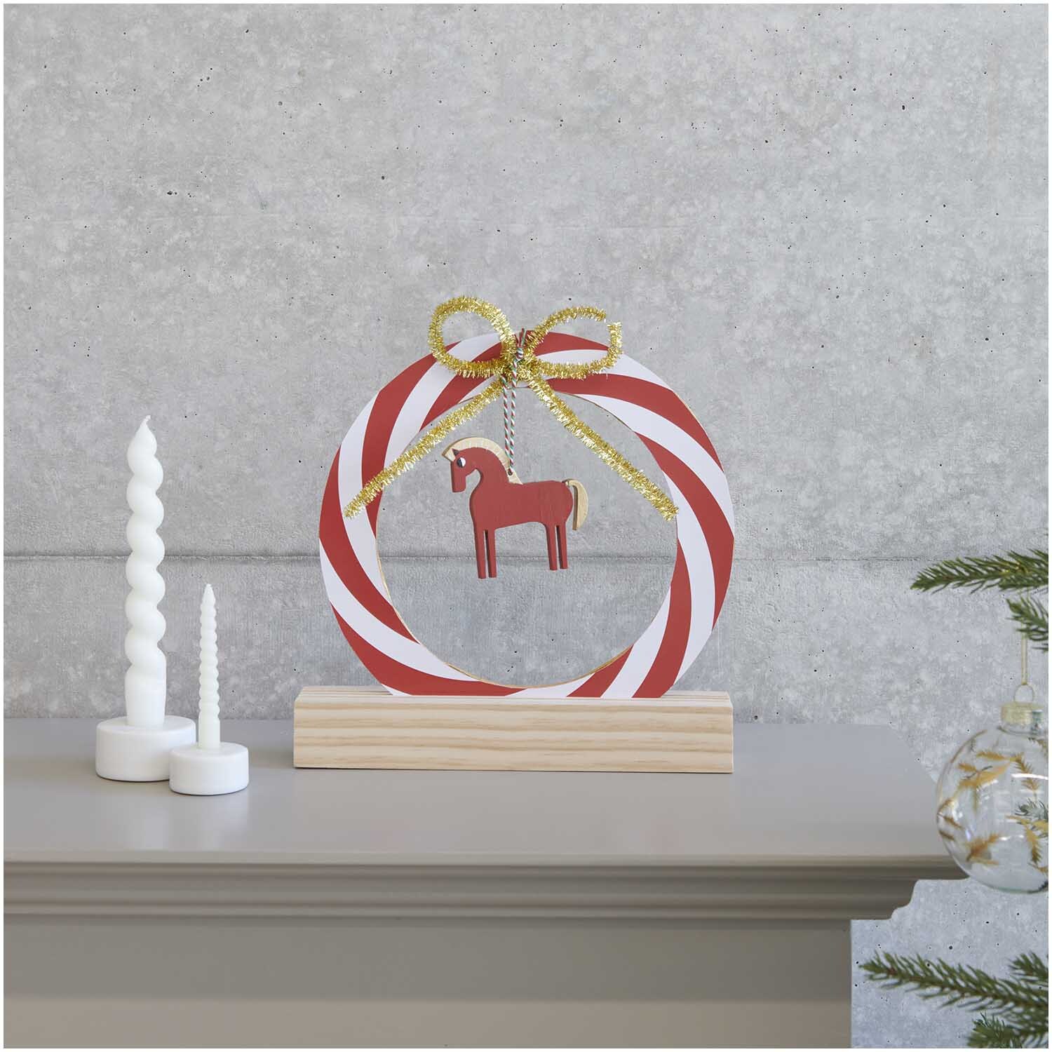 Paper Poetry Kartonring Candy Cane Ø 25cm von Rico Design Paper Poetry Kartonring Candy Cane Ø 25cm mit rot-weißen Streifen und goldener Schleife, dekoriert mit einem Holzpferd, auf einem Holzsockel.