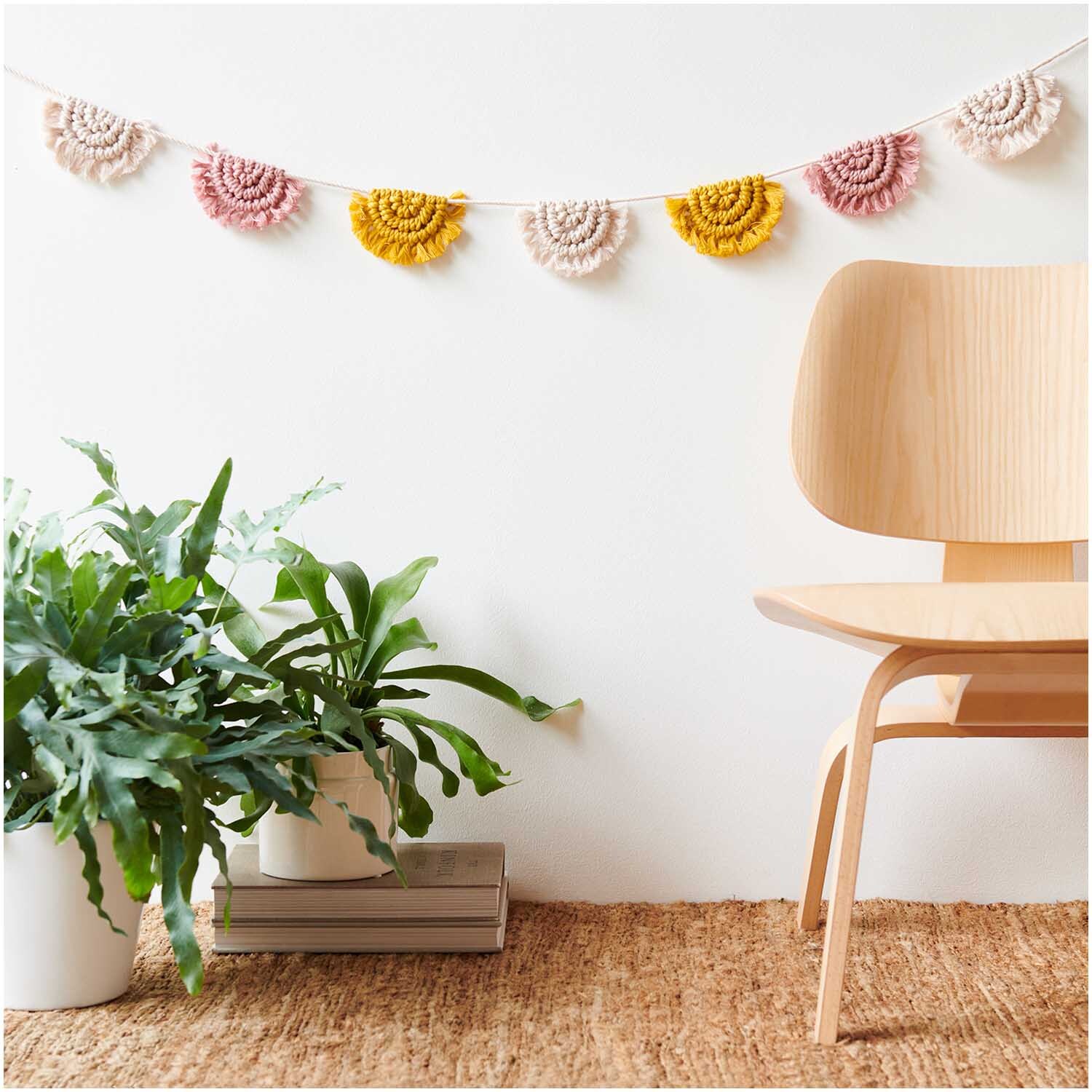 Creative Cotton Cord Makramee-Garn in Senf von Rico Design, dekorativ als Girlande an einer Wand, neben einem Holzstuhl und Pflanzen arrangiert.