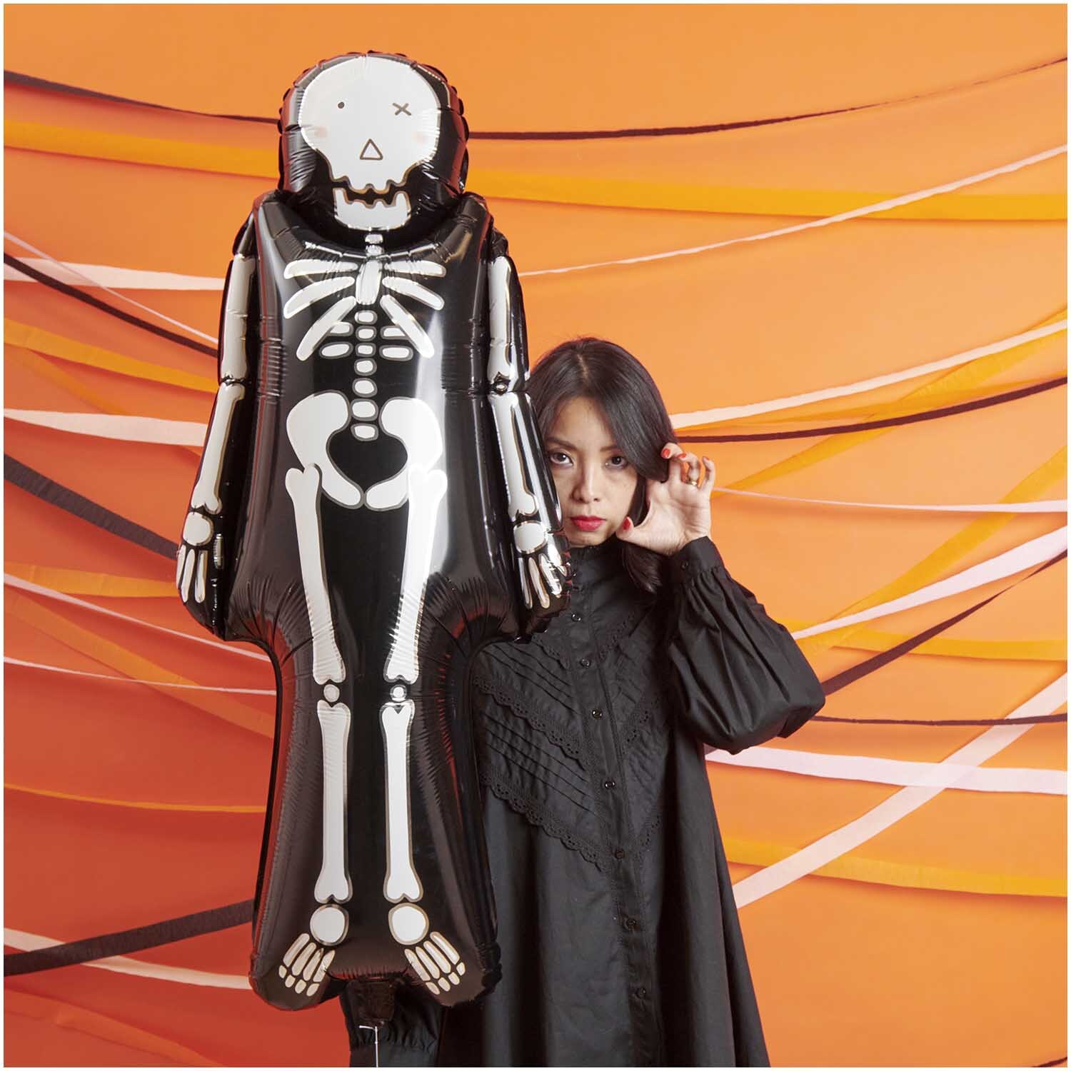 Folienballon Skelett 110x48cm von Rico Design Folienballon Skelett 110x48cm von Rico Design vor orangefarbenem Hintergrund mit Frau in Schwarz.