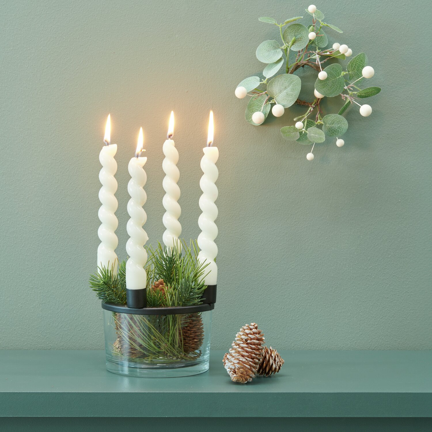 Blätterkranz mit Schneebeeren: Advent-Kerzenhalter mit Spiralkerzen und Fichtenzapfen auf grünem Tisch.