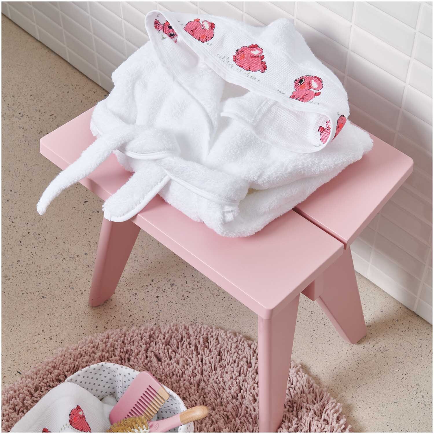 Children's bathrobe from 18 months colour White from Rico Design Kinderbademantel ab 18 Monate auf rosa Hocker, mit Kapuze und niedlichen roten Motiven. Ideal für Kleinkinder.