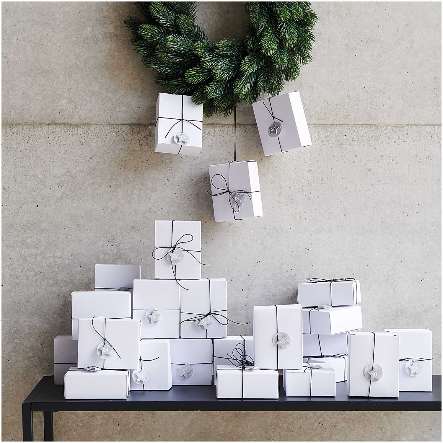 Holzsticker Adventskalender Sterne 1-24: Weiße Geschenke mit schwarzem Band unter einem grünen Kranz, dekoriert für die Adventszeit.