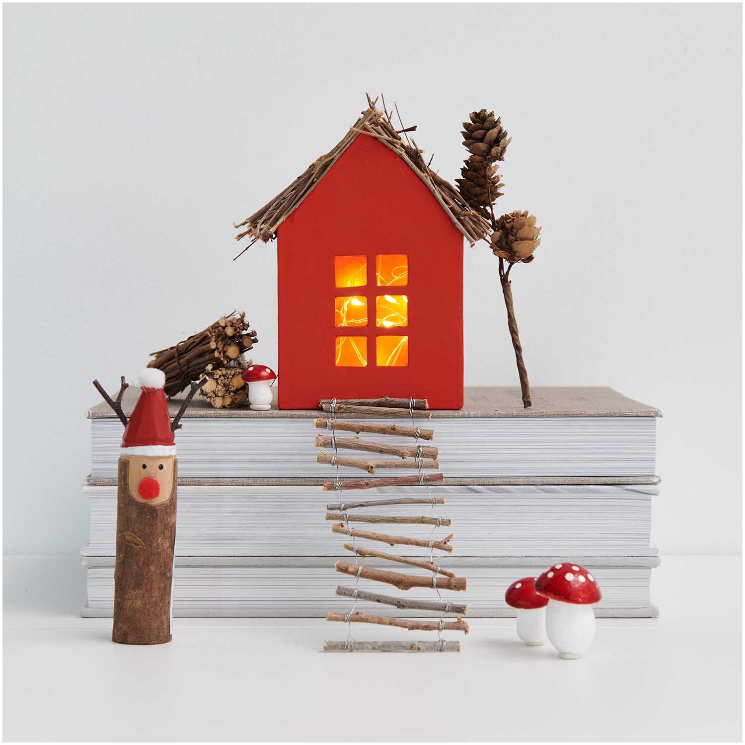 Holzfigur Rentier 7x12,5cm von Rico Design Holzfigur Rentier 7x12,5cm von Rico Design vor einem roten Haus mit leuchtenden Fenstern und Naturdeko.