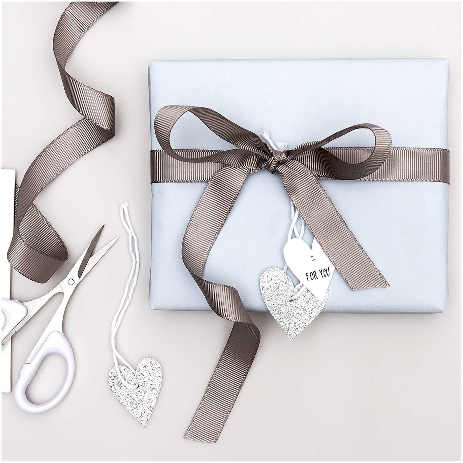 Paper Poetry Papieranhänger Herzen 3x3,5cm Glitter silber 24 Stück von Rico Design Paper Poetry Papieranhänger Herzen 3x3,5cm Glitter silber 24 Stück, dekorativ an Geschenk mit braunem Band, neben Schere und weiteren Anhängern.