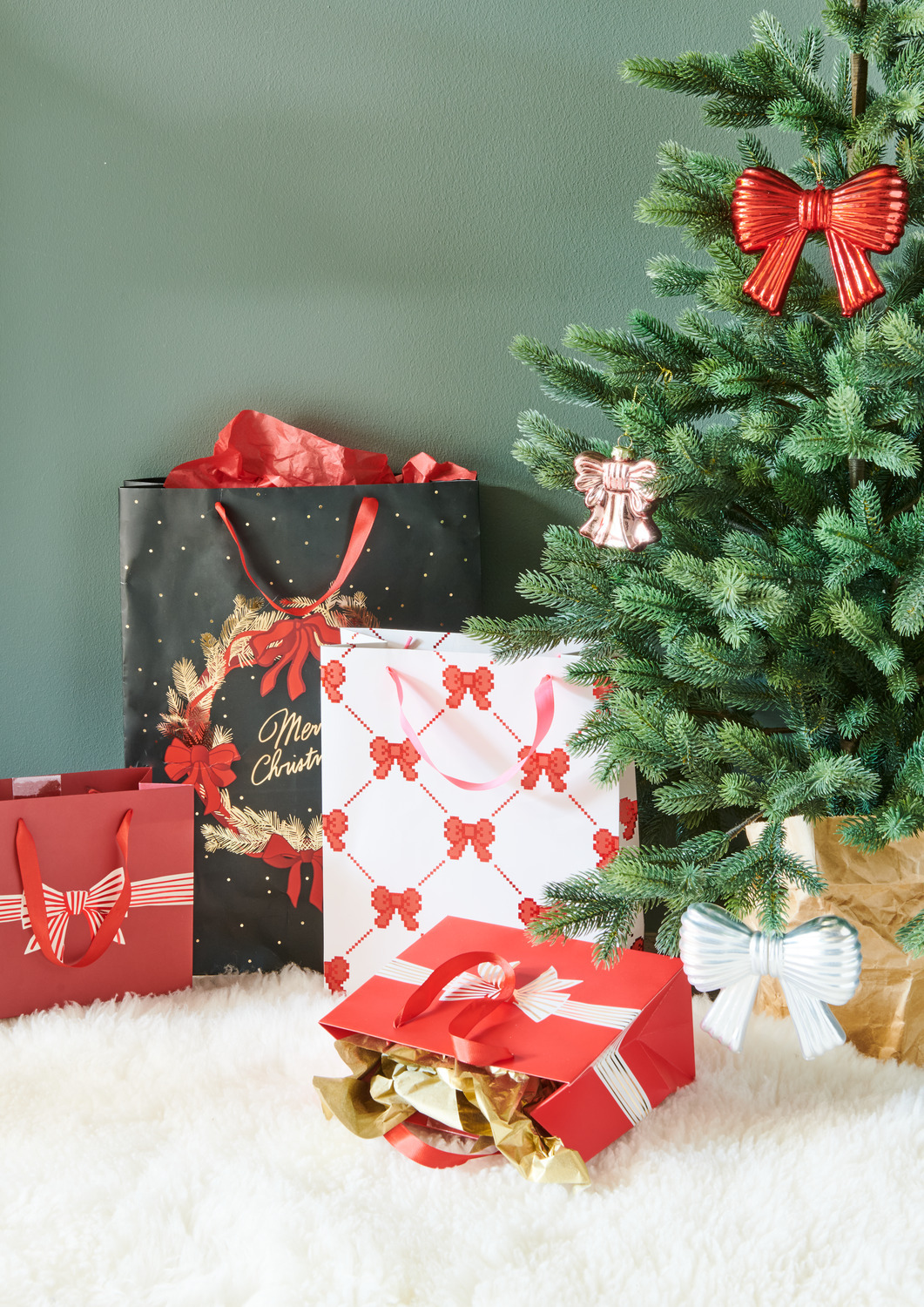Geschenktüte Pixel Schleifen Creme L 26x32x12cm, 230g/m² von Rico Design Geschenktüte Pixel Schleifen Creme L neben einem Weihnachtsbaum mit roten und weißen Schleifen, umgeben von weiteren festlichen Geschenktüten.