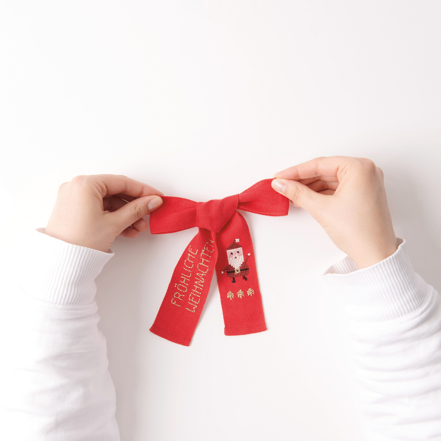 Stickpackung Schleife Nikolaus 12x19cm von Rico Design Stickbuch Put a Bow on It Nr. 185: Rote Schleife mit Nikolausmotiv und Schriftzug "Fröhliche Weihnachten", gehalten von Händen.