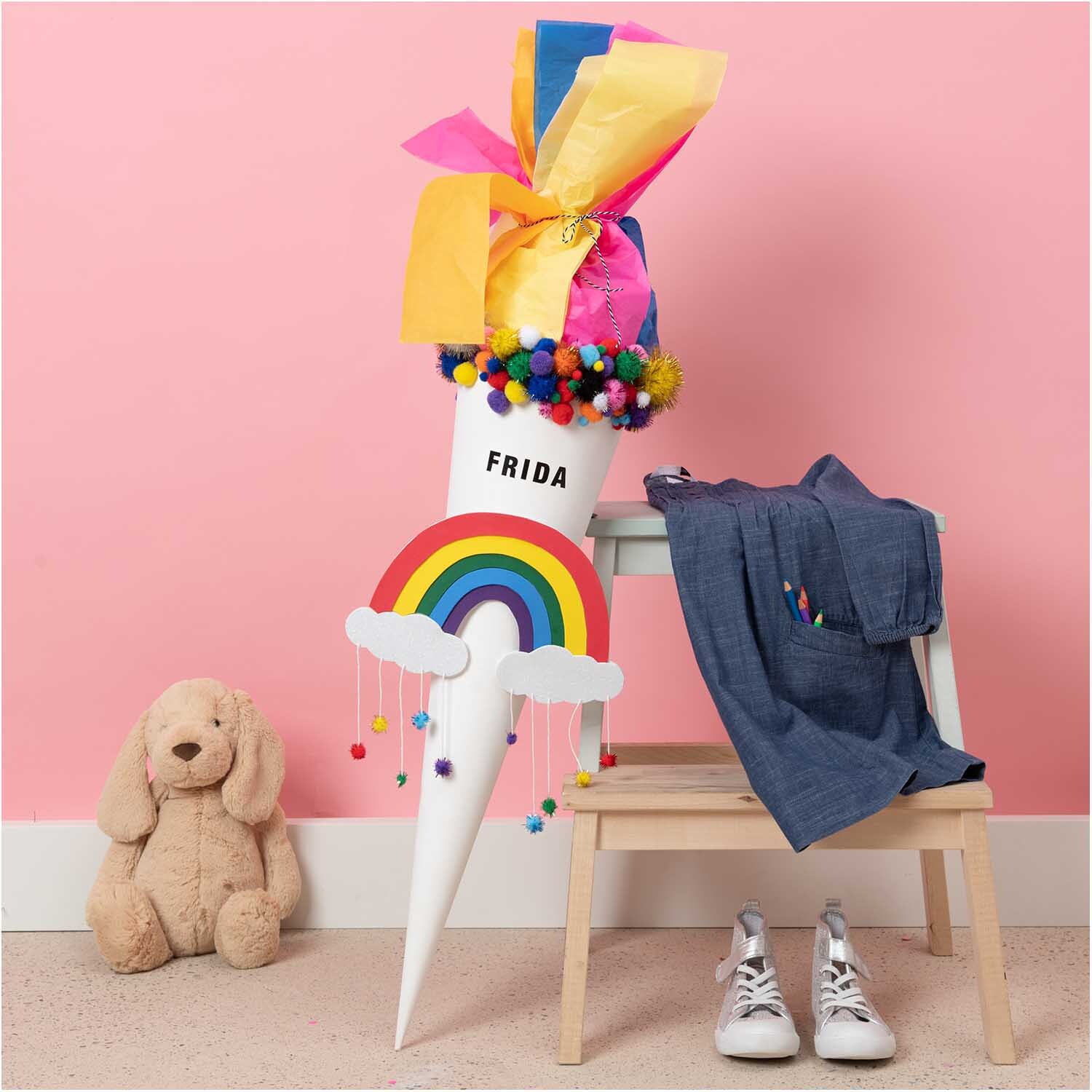 Glitter Pompons 63 Stück Farbe Bunt von Rico Design Glitter Pompons 63 Stück von Rico Design in einer bunten Schultüte mit Regenbogenmotiv, umgeben von Kleidung und einem Plüschtier.