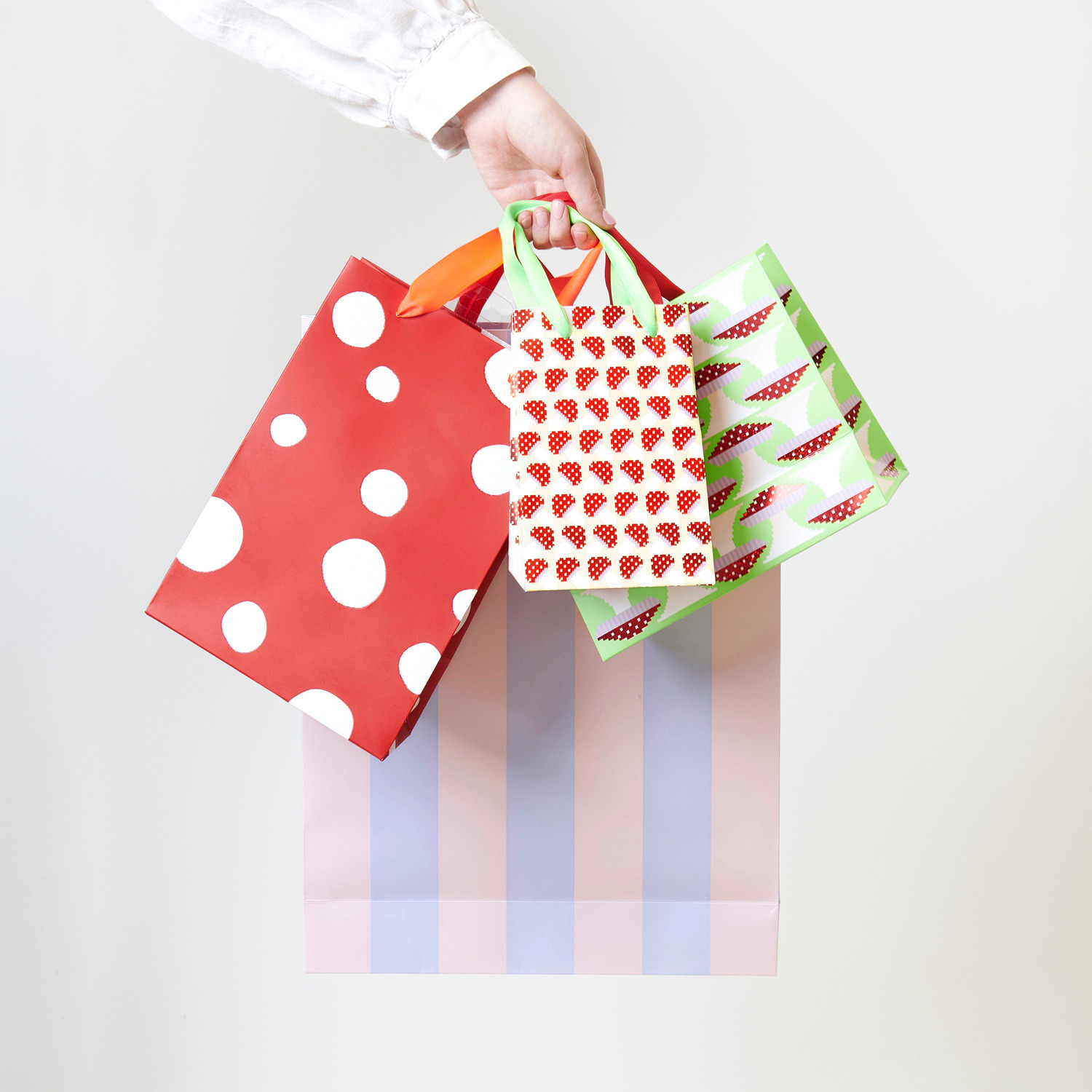 Geschenktüte Streifen Rosa-Flieder 18x26x12cm von Rico Design Geschenktüte Fliegenpilz Pixel Grün, gehalten von einer Hand, mit bunten Mustern und Streifen.